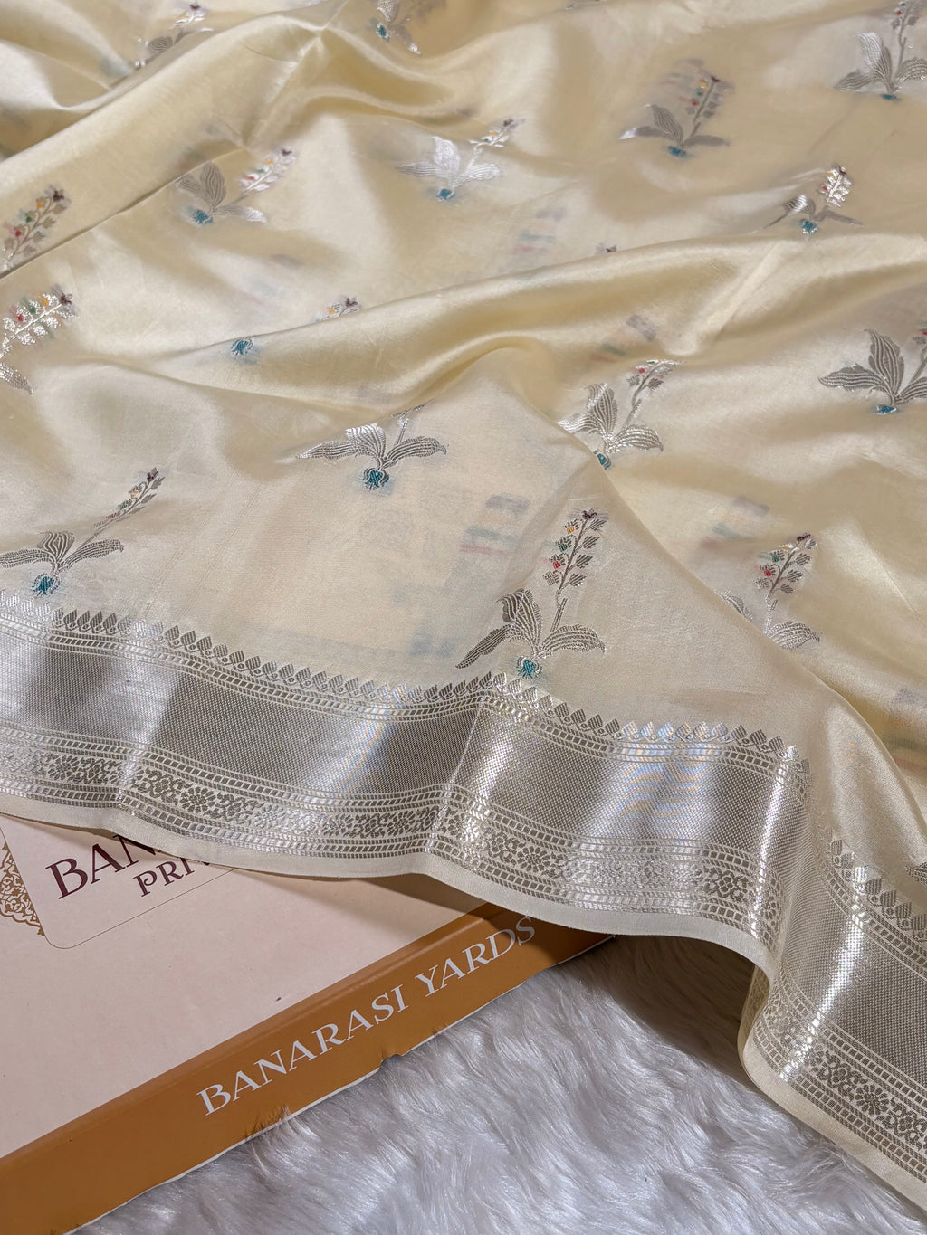 A close-up of a cream-colored Banarasi silk saree with silver floral weaving and a wide silver border, draped over a book labeled 'BANARASI YARDS' on a white faux fur surface.