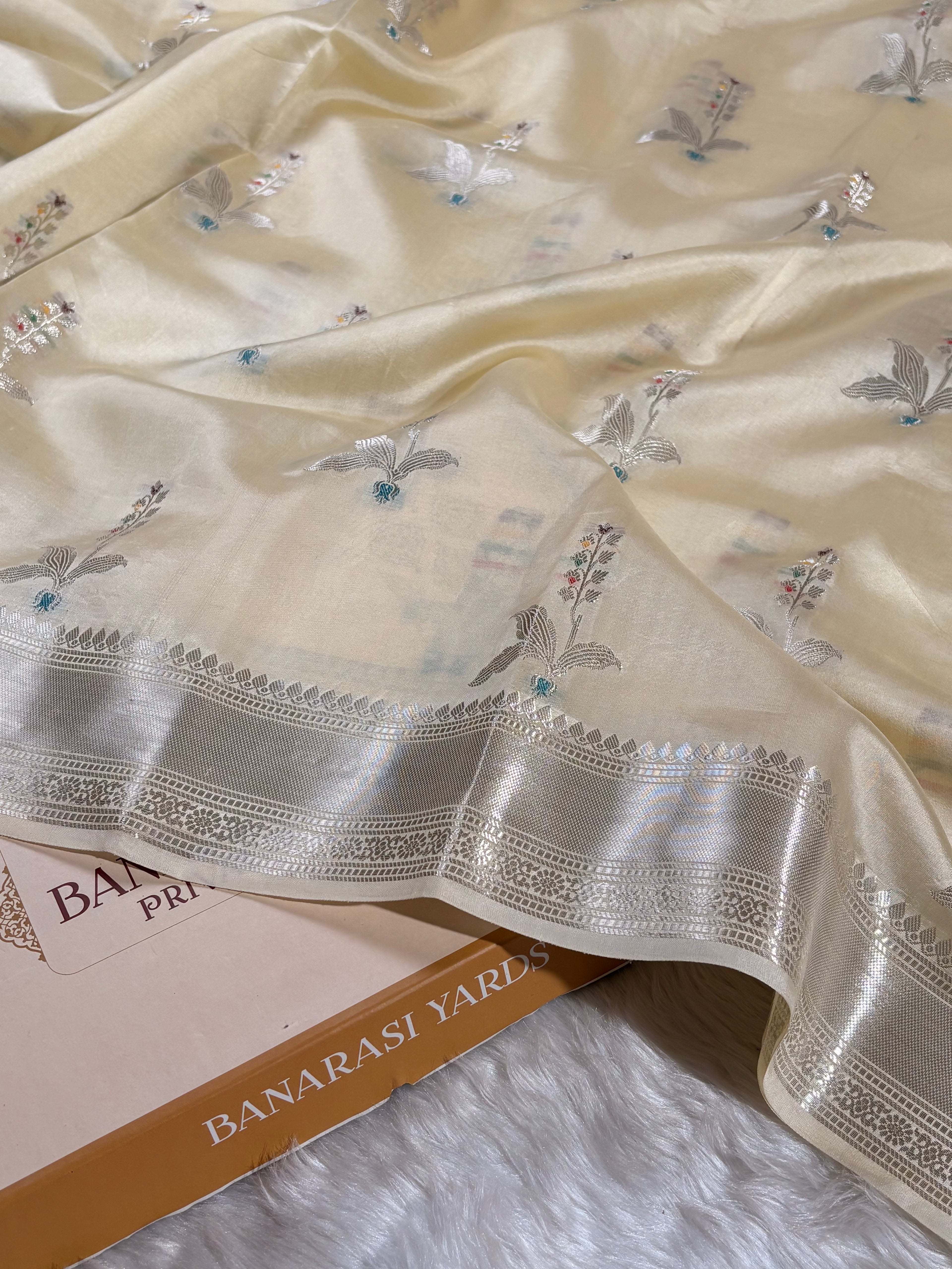 A close-up of a cream-colored Banarasi silk saree with silver floral weaving and a wide silver border, draped over a book labeled 'BANARASI YARDS' on a white faux fur surface.
