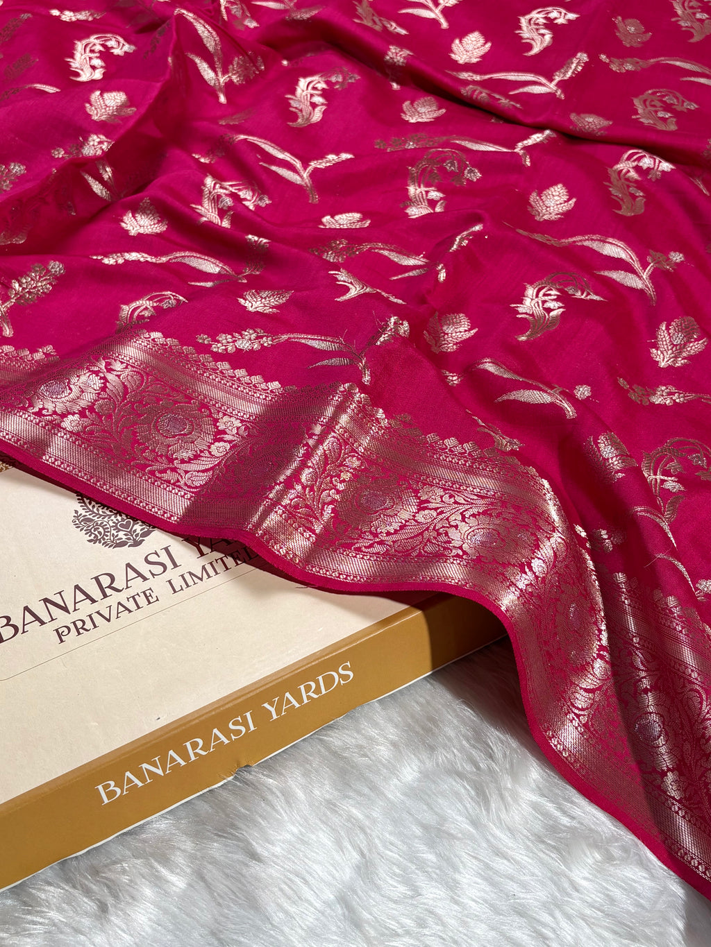 A close-up of a rich magenta pink Banarasi silk saree with intricate gold zari floral motifs and a wide decorative, draped over a Banarasi Yards box on a white faux fur surface.