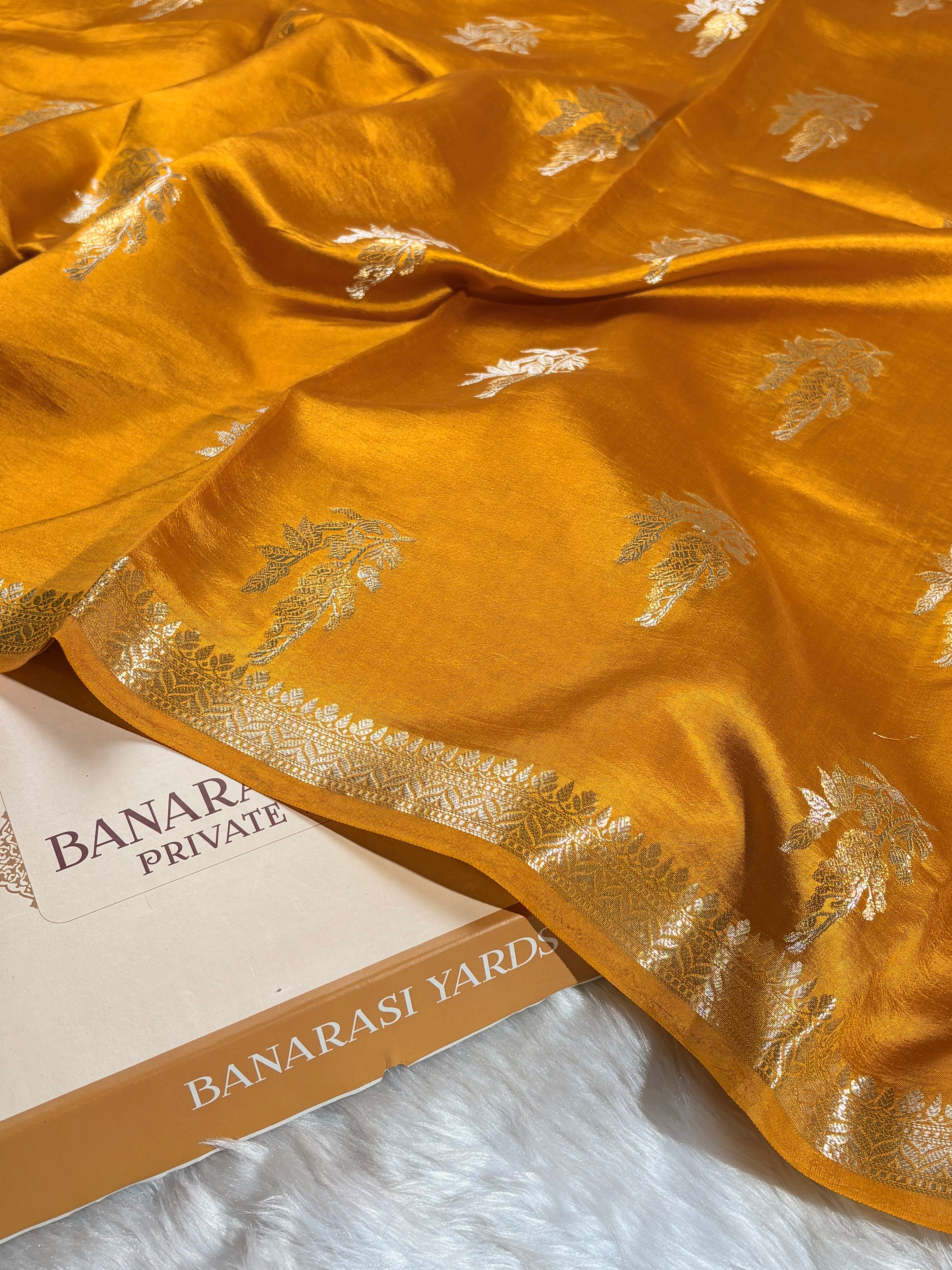 A close-up of a mustard yellow Banarasi silk saree with silver zari floral motifs and a decorative border, draped over a white faux fur surface next to a box labeled 'BANARASI YARDS'.