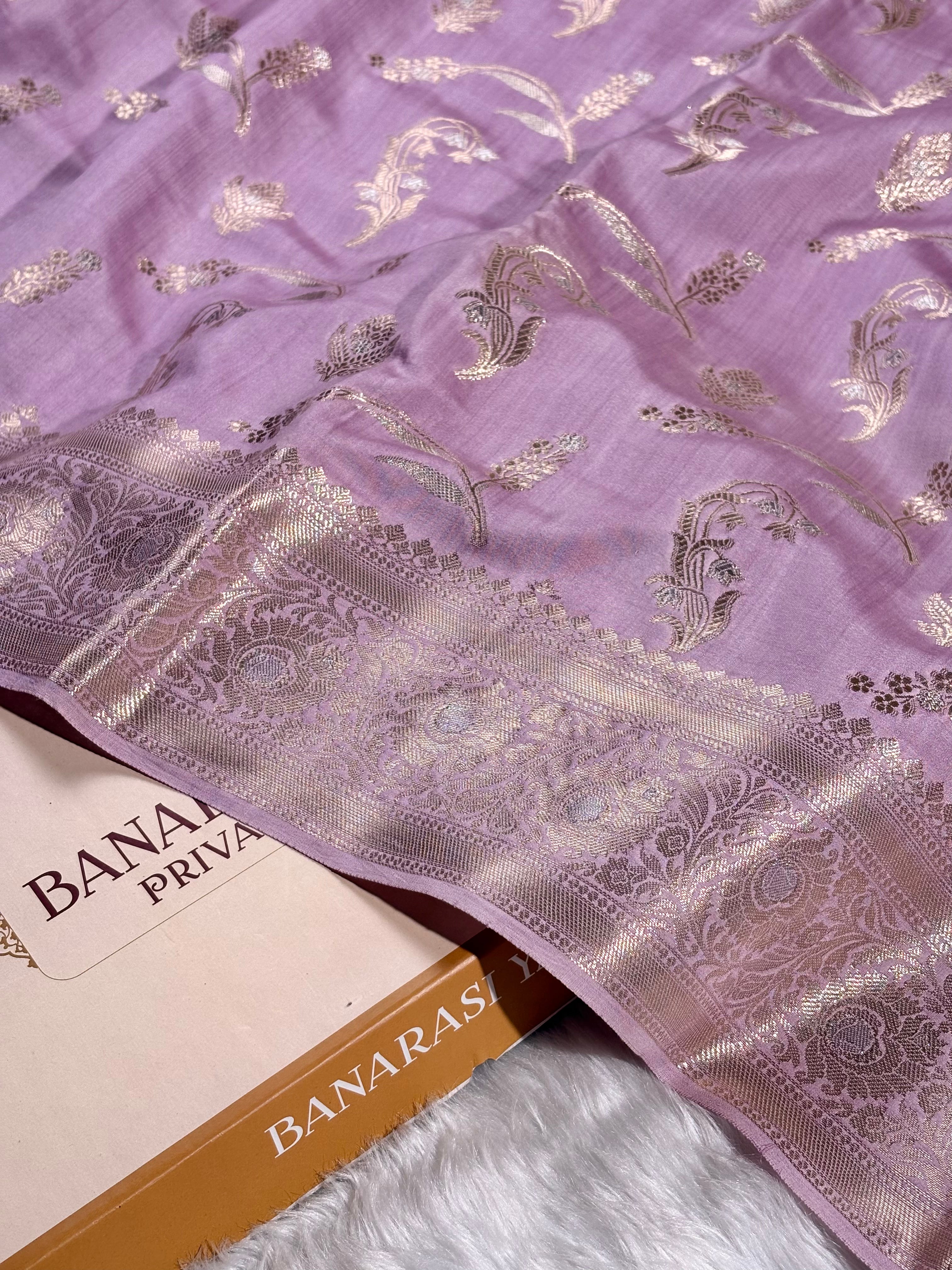 A close-up of a lavender purple Banarasi silk saree with metallic gold floral weaving and a wide patterned border, draped over a book labeled 'BANARASI YARDS' on a white faux fur surface.