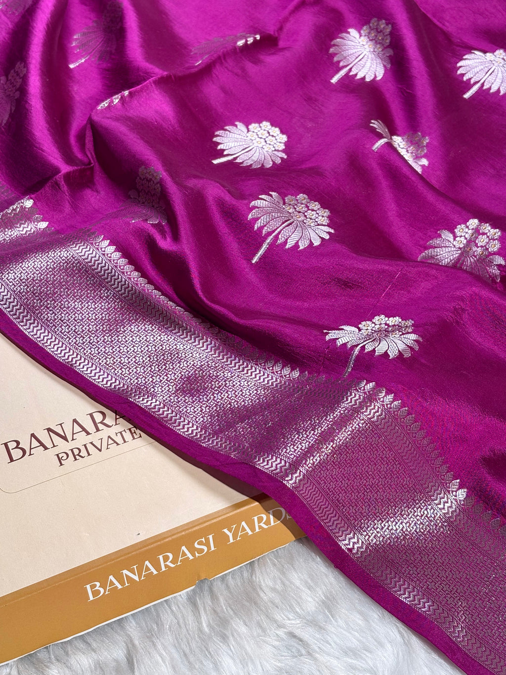 A close-up of a rich magenta purple Banarasi silk saree with silver floral motifs and a detailed zari border, draped over a white faux fur surface next to a paper label reading 'BANARASI YARDS'.