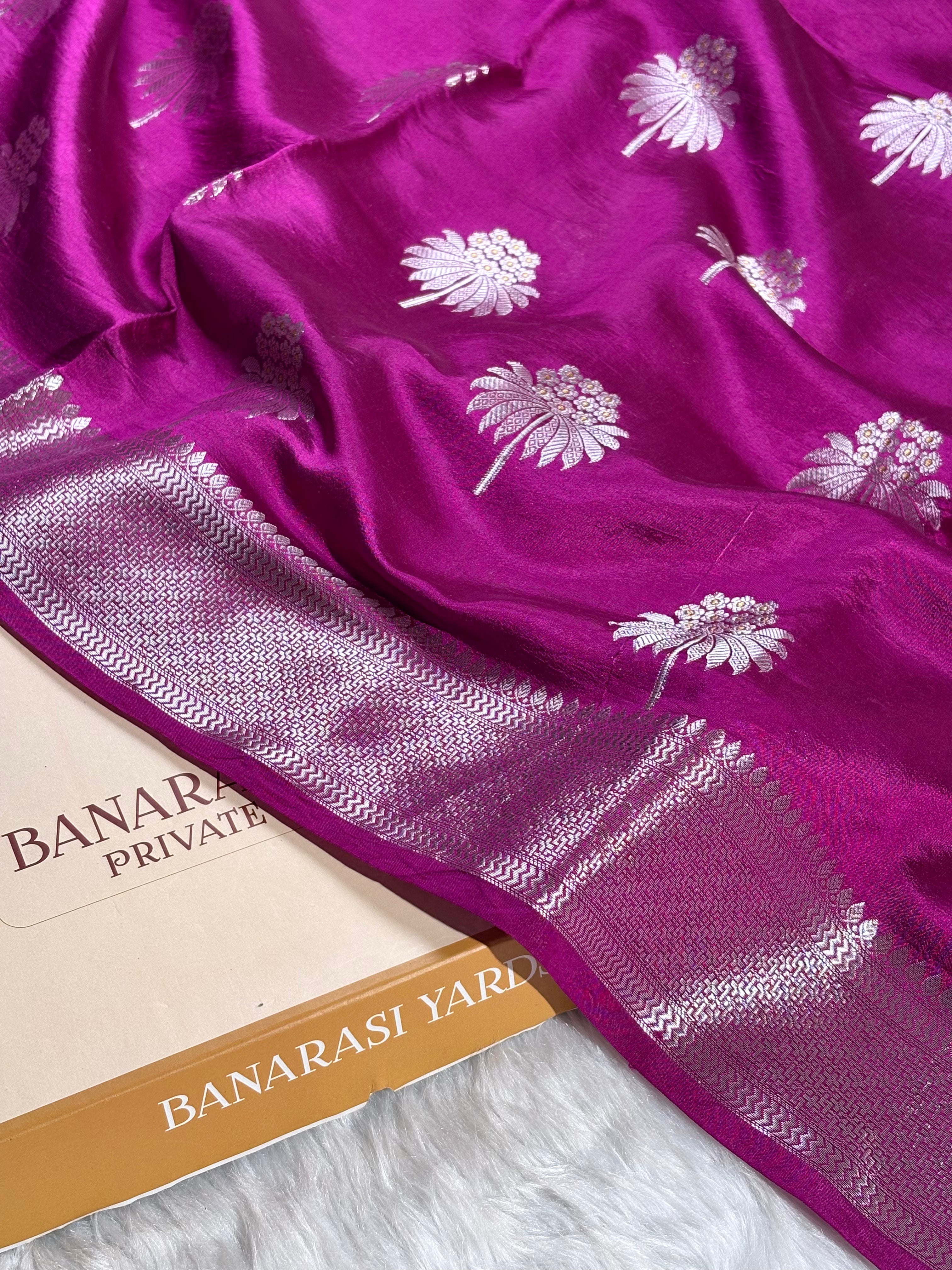 A close-up of a rich magenta purple Banarasi silk saree with silver floral motifs and a detailed zari border, draped over a white faux fur surface next to a paper label reading 'BANARASI YARDS'.