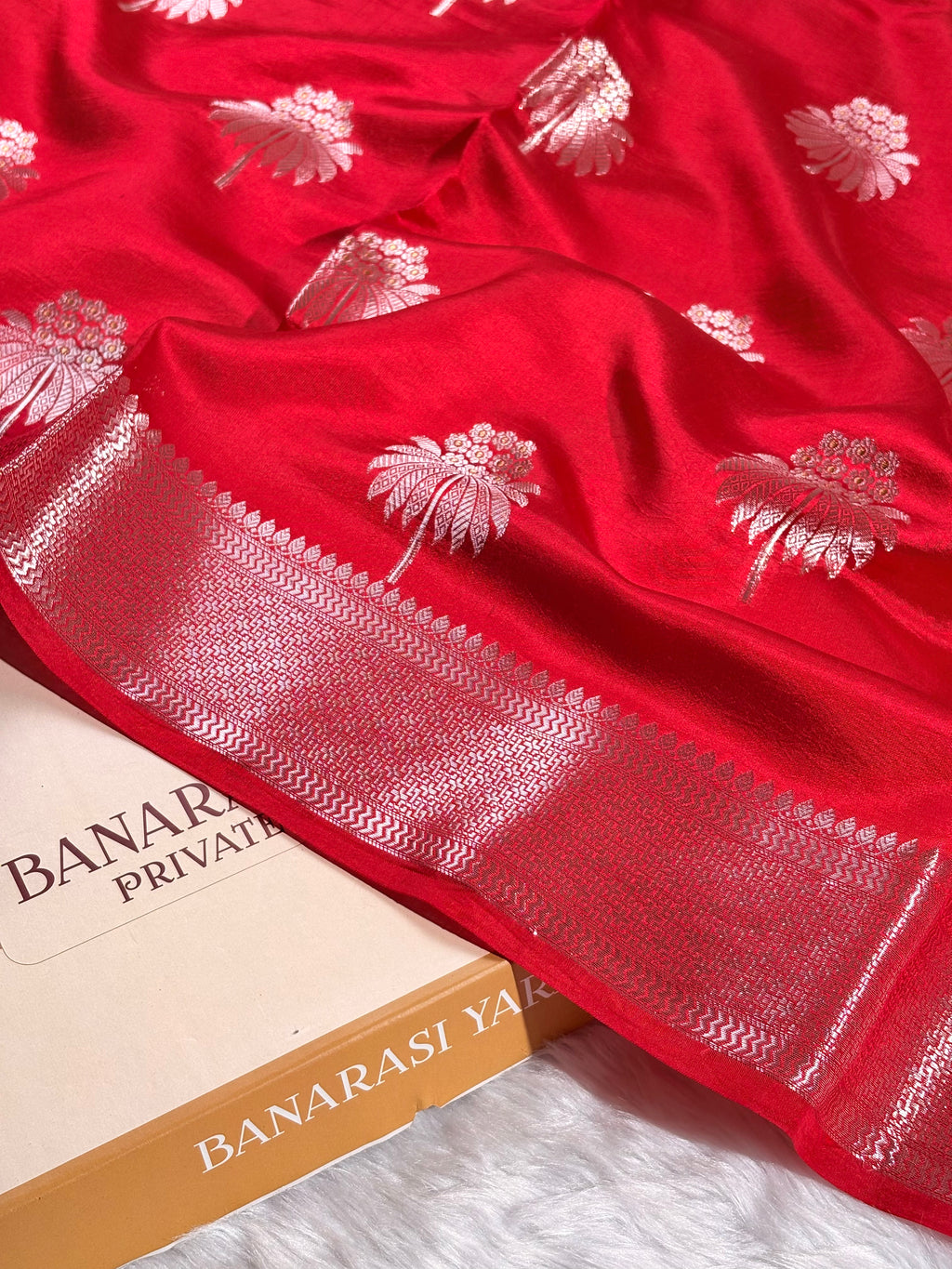 A close-up of a bright red Banarasi silk saree with alfi mina floral motifs and a wide zari border, draped over a Banarasi Yards box on a white faux fur surface.