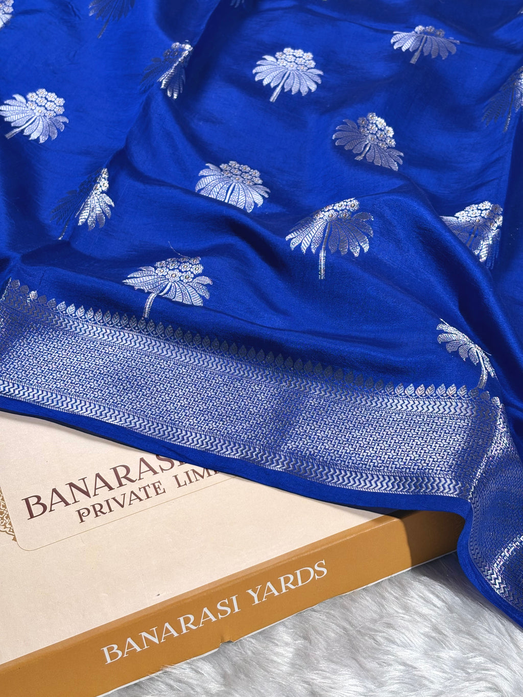 A royal blue Banarasi silk saree with alfi mina weaving and a detailed zari border, draped over a Banarasi Yards box on a white textured surface.