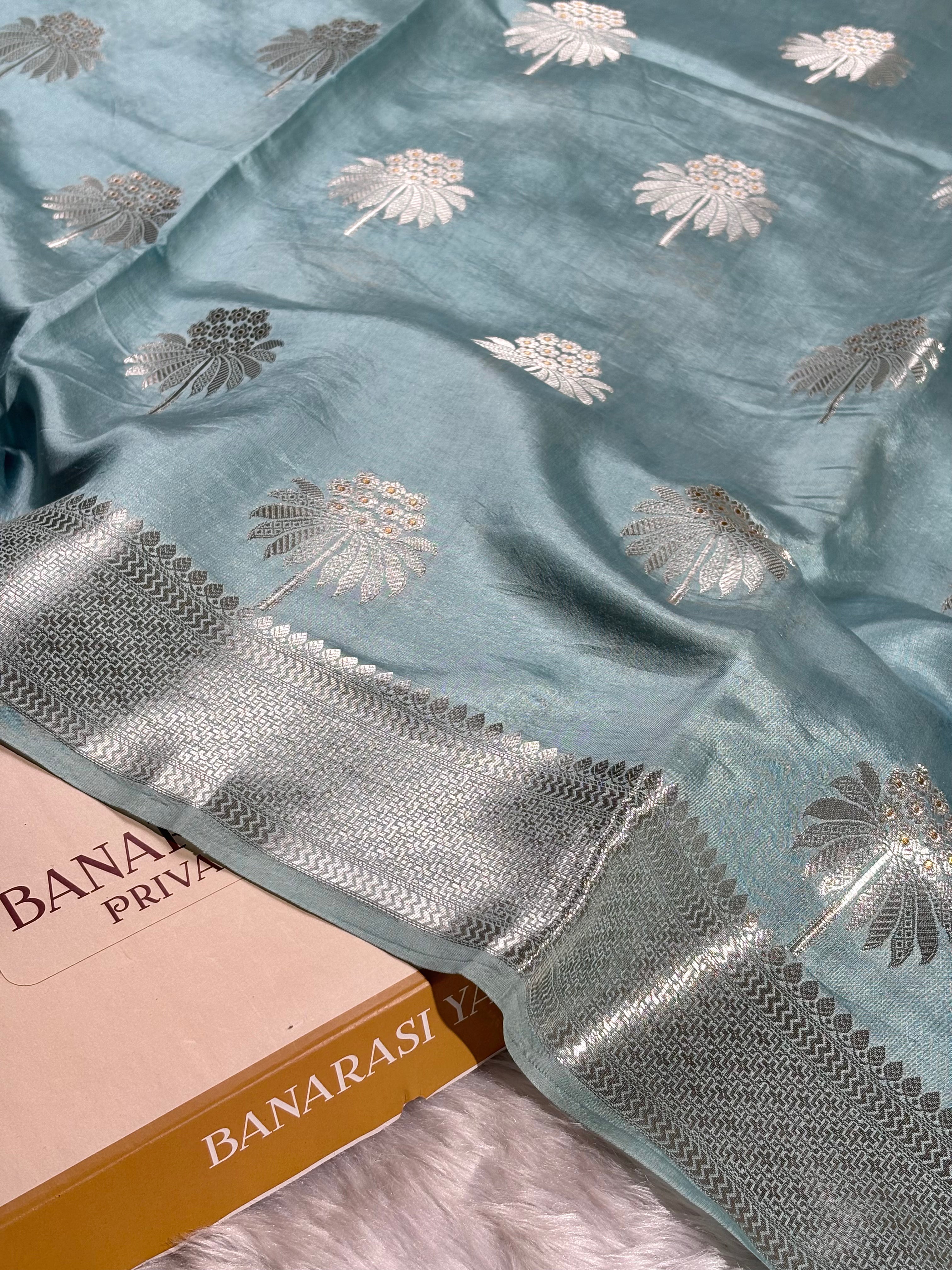 A light blue-green Banarasi silk saree with silver floral motifs and a wide silver border, styled on a white faux fur surface next to a Banarasi Yards box.