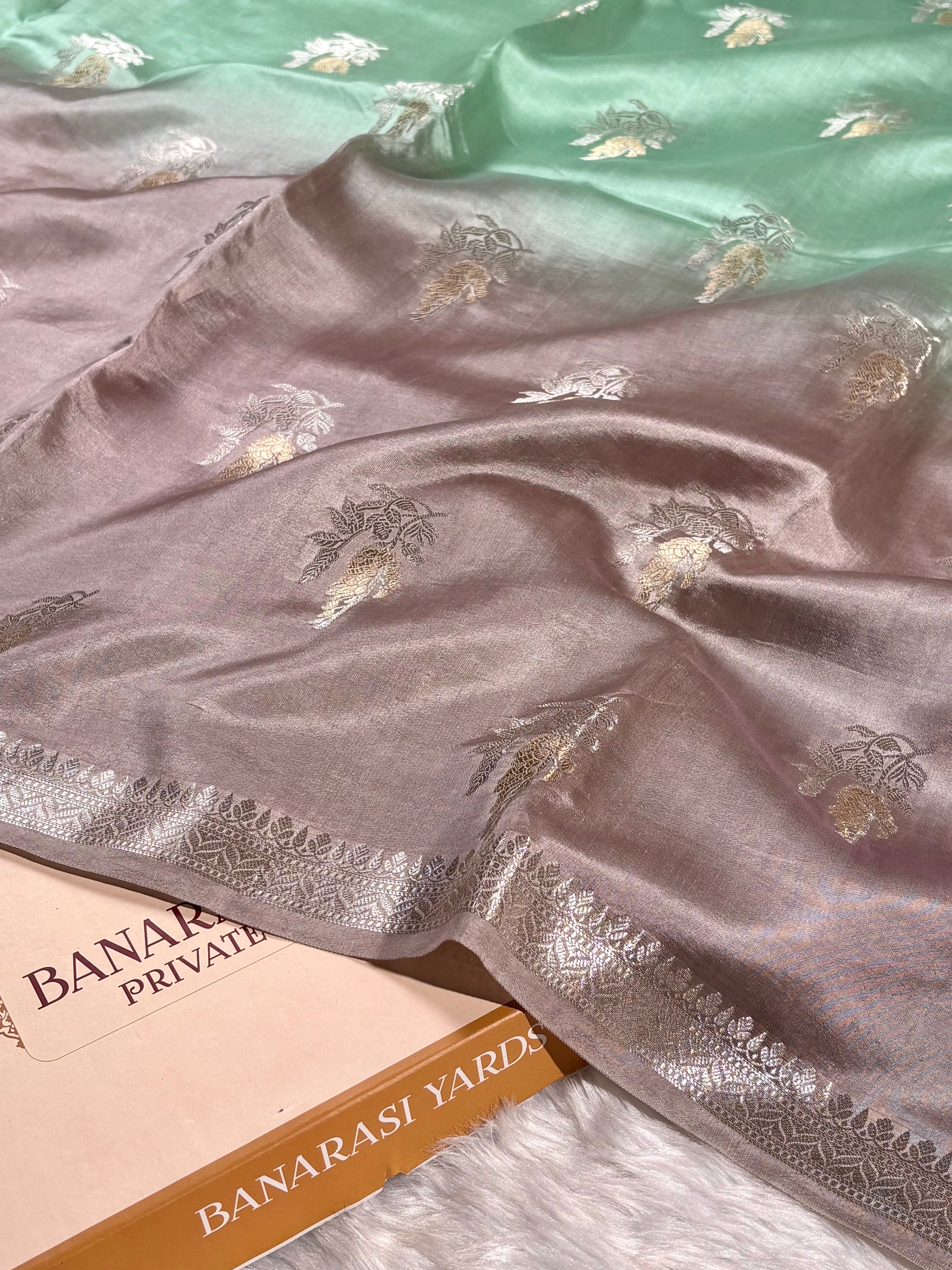 A Banarasi silk saree draped to show its dual-color design, transitioning from a dusty mauve-pink lower body with alfi mina floral zari weaving to a mint green upper body. A box branded 'BANARASI YARDS' is partially visible underneath.