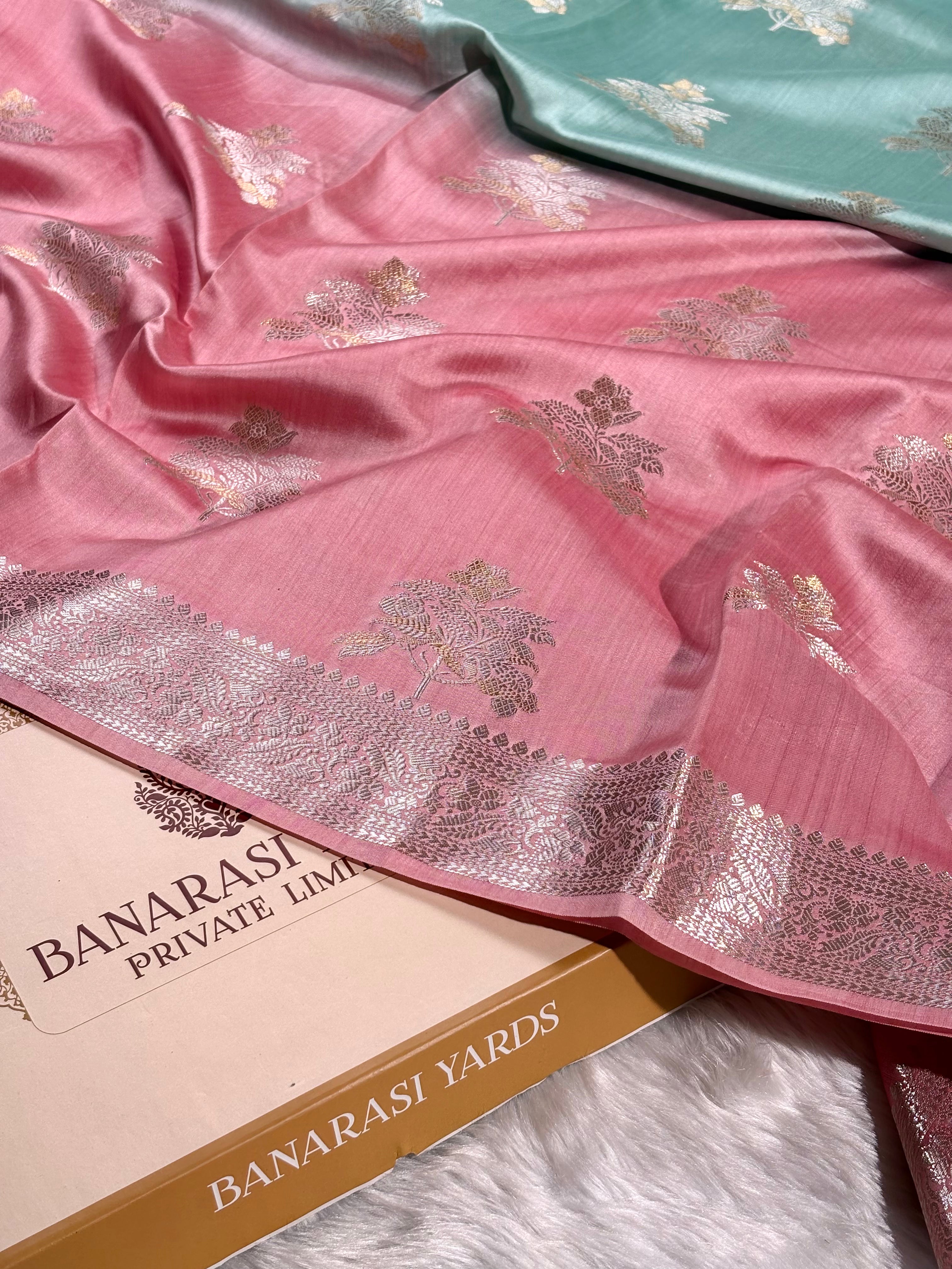 Pink Banarasi silk saree with gold floral motifs and a wide alfi mina zari border, draped over a Banarasi Yards Private Limited box on a white faux fur surface.