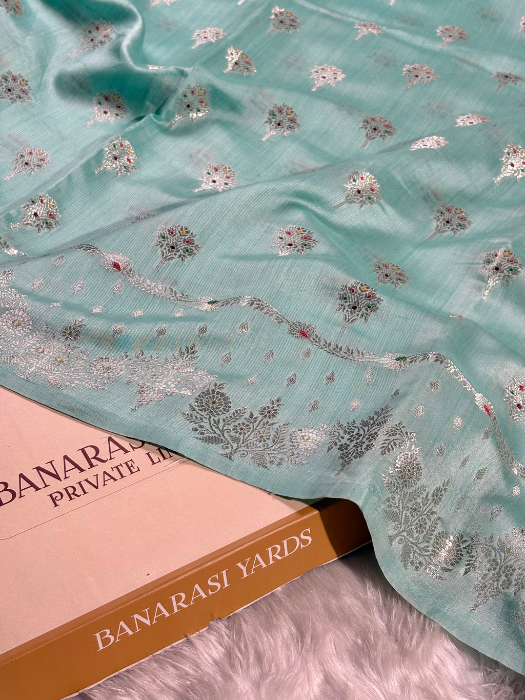 A mint green Banarasi silk saree, decorated with tilfi mina floral motifs and a zari border, draped over a labeled 'BANARASI YARDS' on a white faux fur surface.