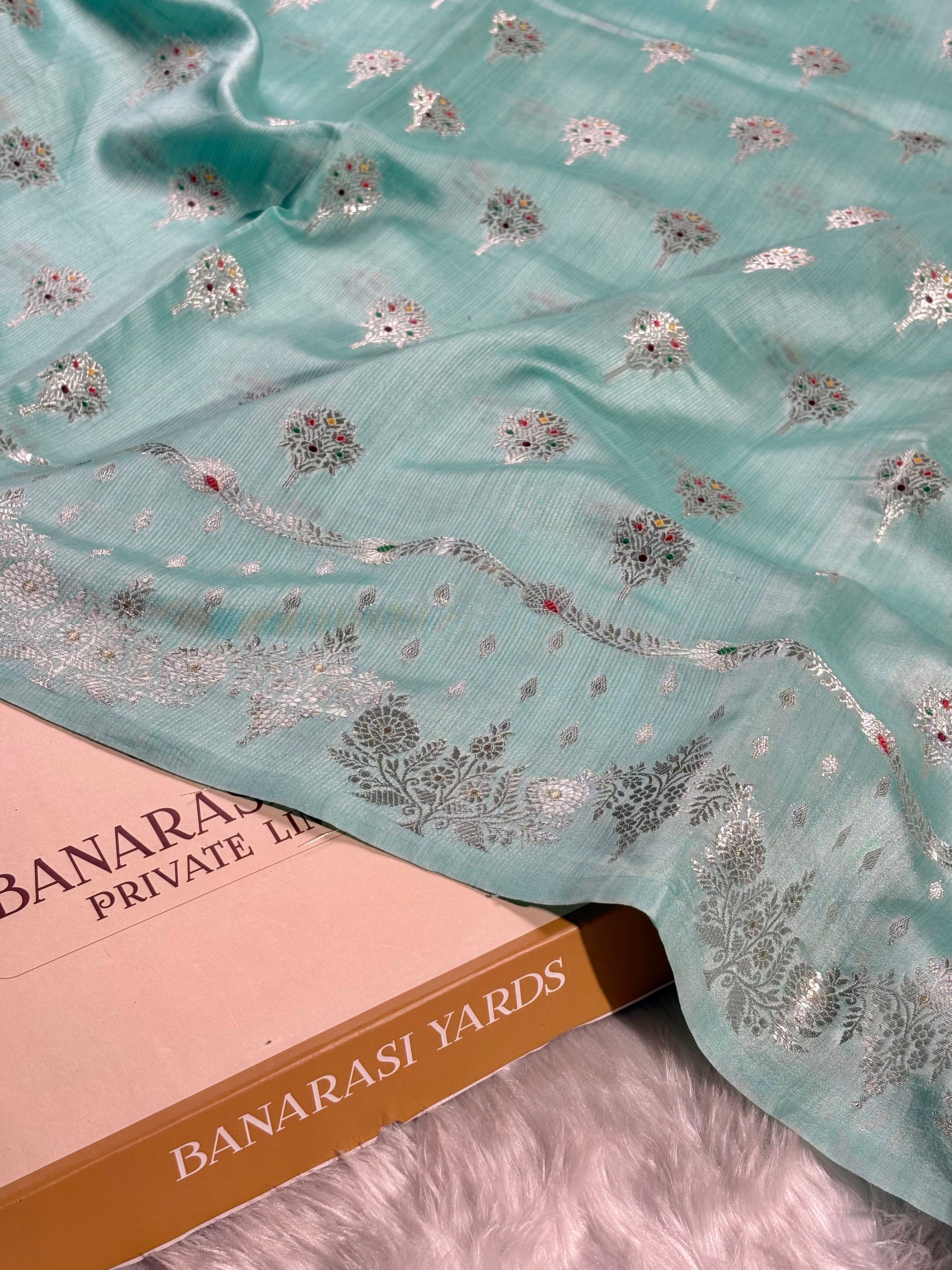 A mint green Banarasi silk saree, decorated with tilfi mina floral motifs and a zari border, draped over a labeled 'BANARASI YARDS' on a white faux fur surface.