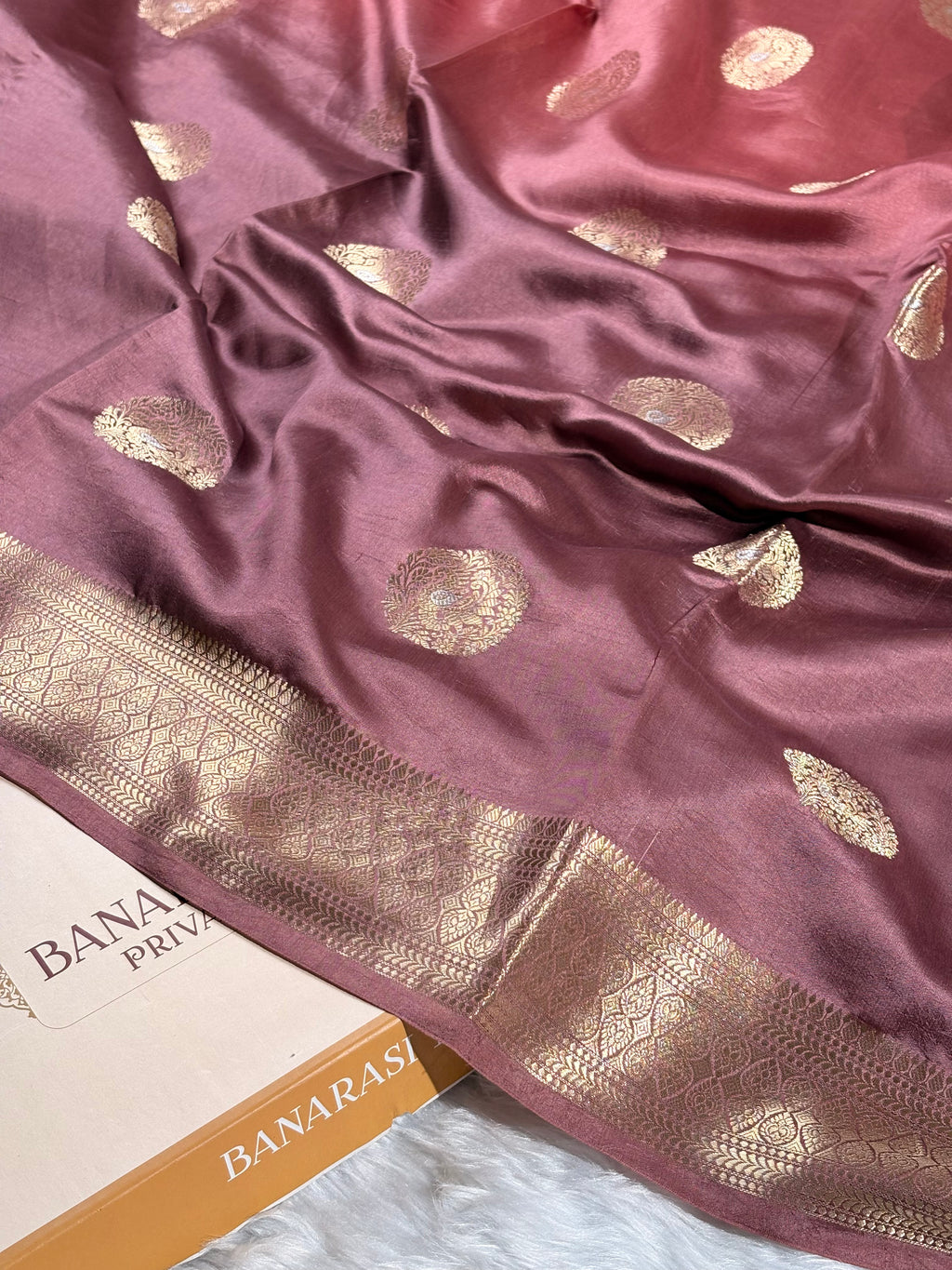 A close-up of a dusty rose pink silk saree with gold zari woven motifs and a wide decorative gold border, draped over a white surface with a Banarasi partially visible.