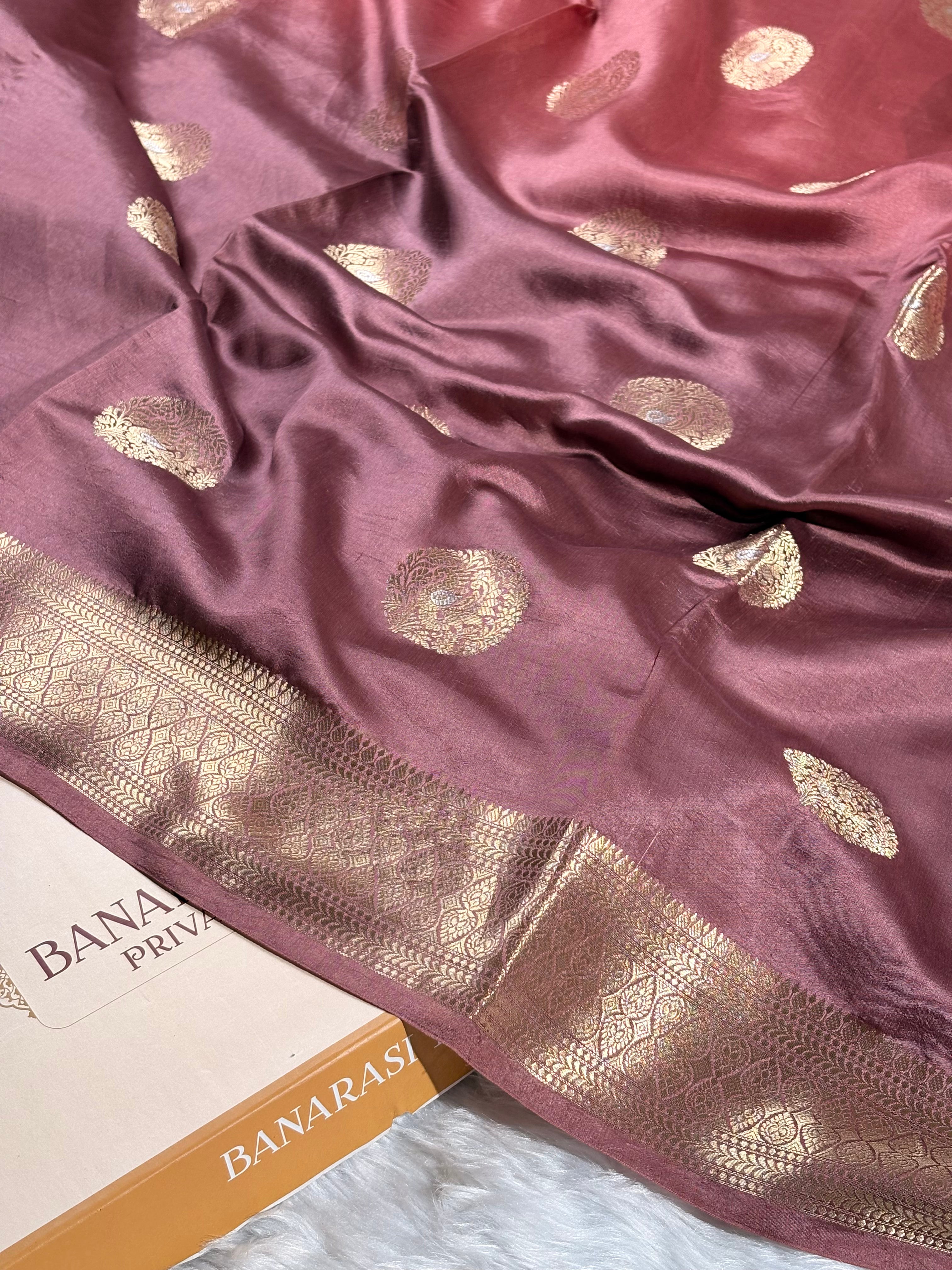 A close-up of a dusty rose pink silk saree with gold zari woven motifs and a wide decorative gold border, draped over a white surface with a Banarasi partially visible.