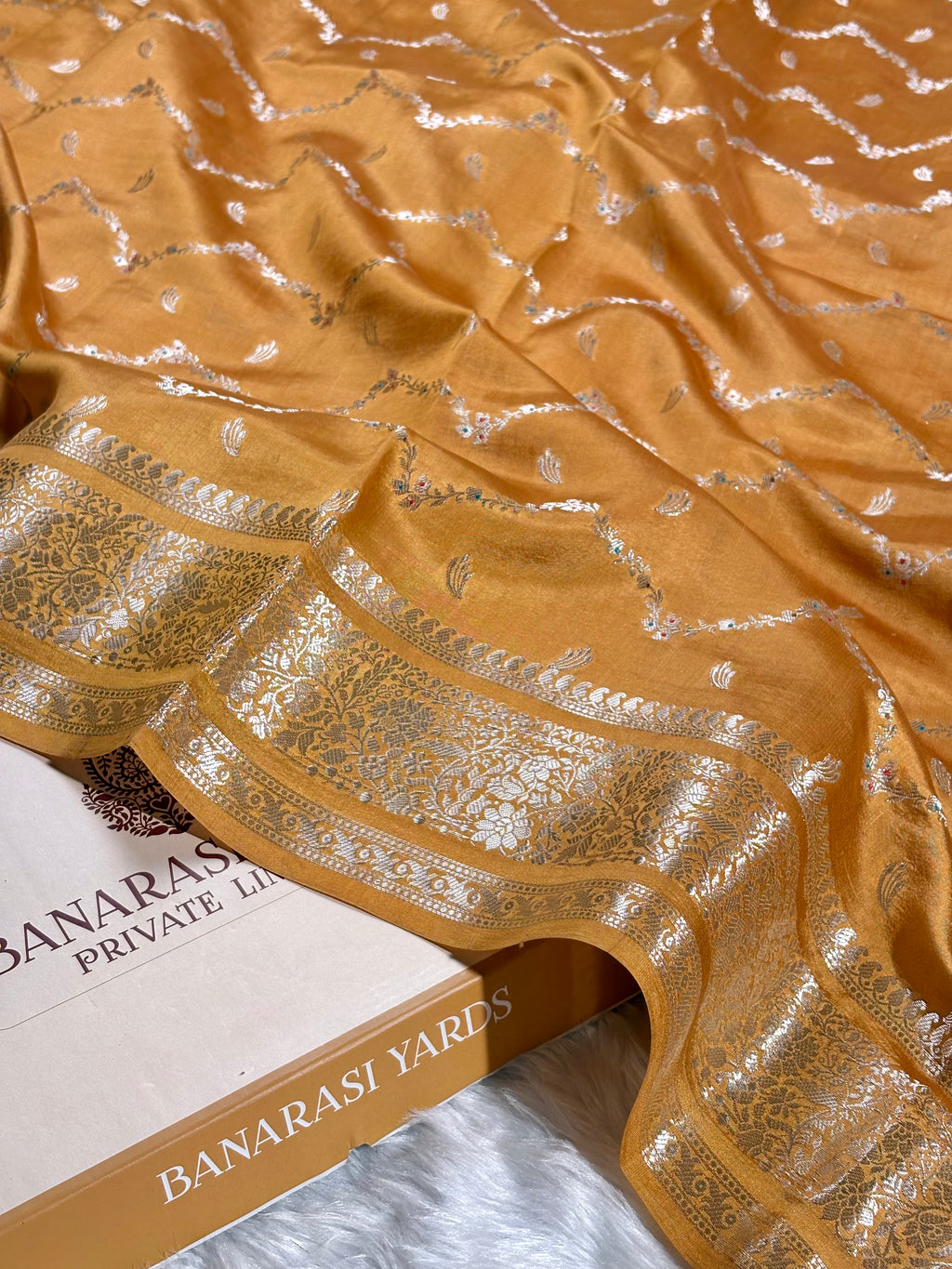 A close-up of a mustard yellow Banarasi silk saree with intricate zari weaving and a wide decorative border, draped over a box labeled 'BANARASI YARDS'.