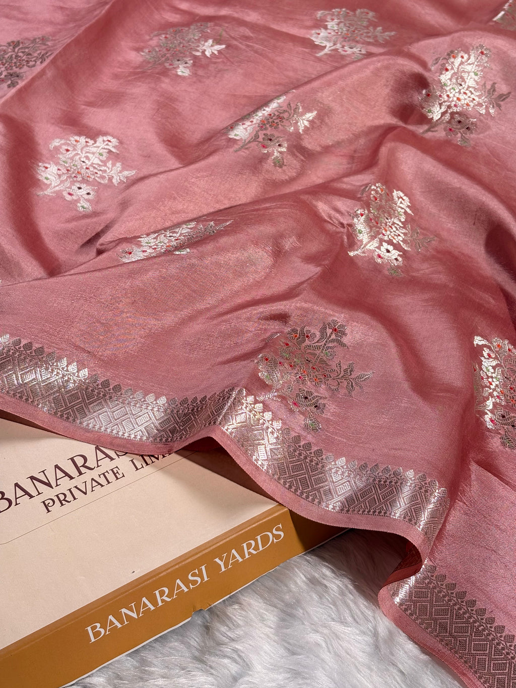 A dusty pink Banarasi silk saree with tilfi mina floral motifs and a patterned border, draped over a book labeled 'BANARASI YARDS' on a white faux fur surface.