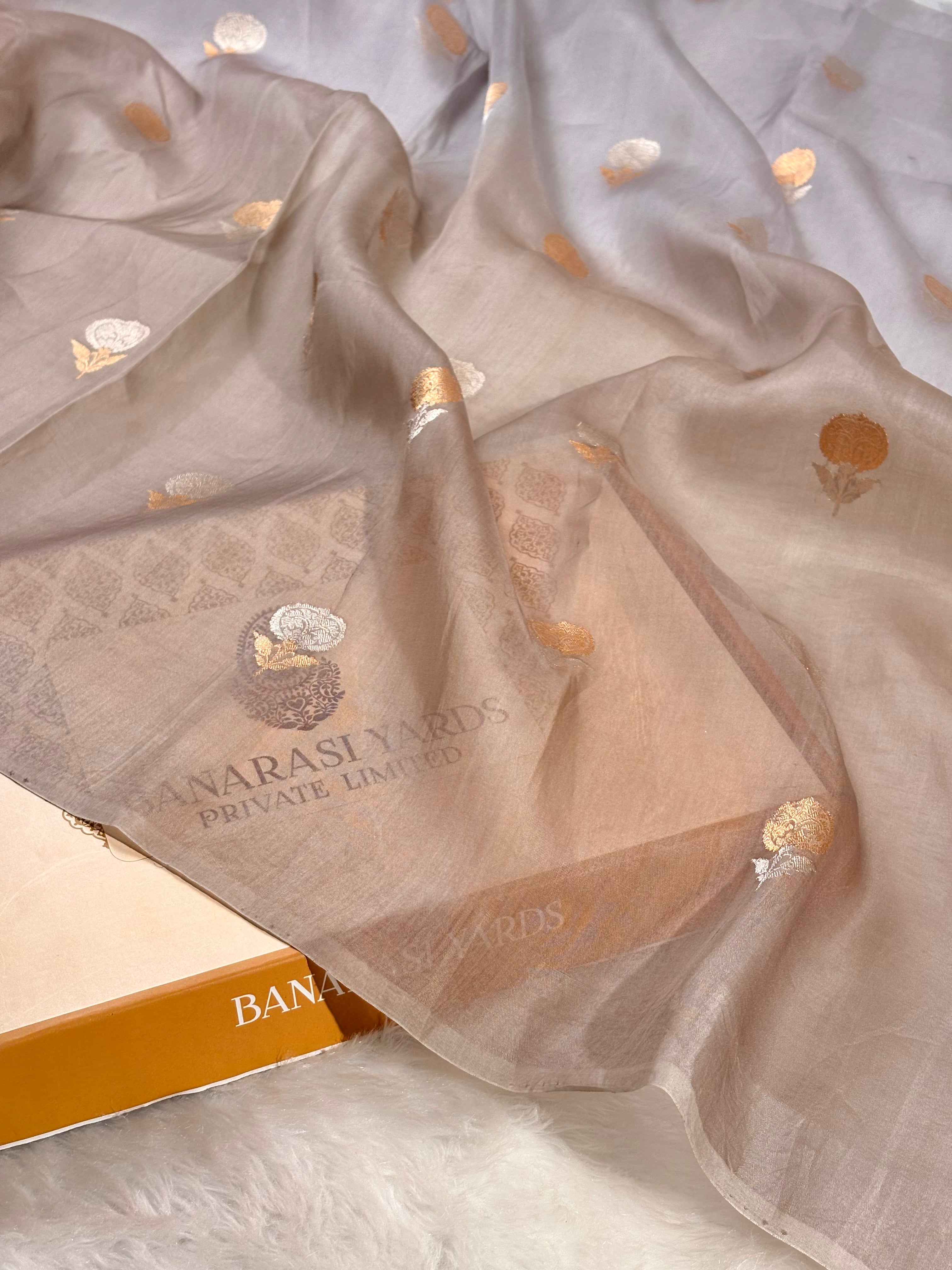 A sheer beige organza dupatta with sona rupa floral motifs draped over a box partially visible underneath on a white faux fur surface.