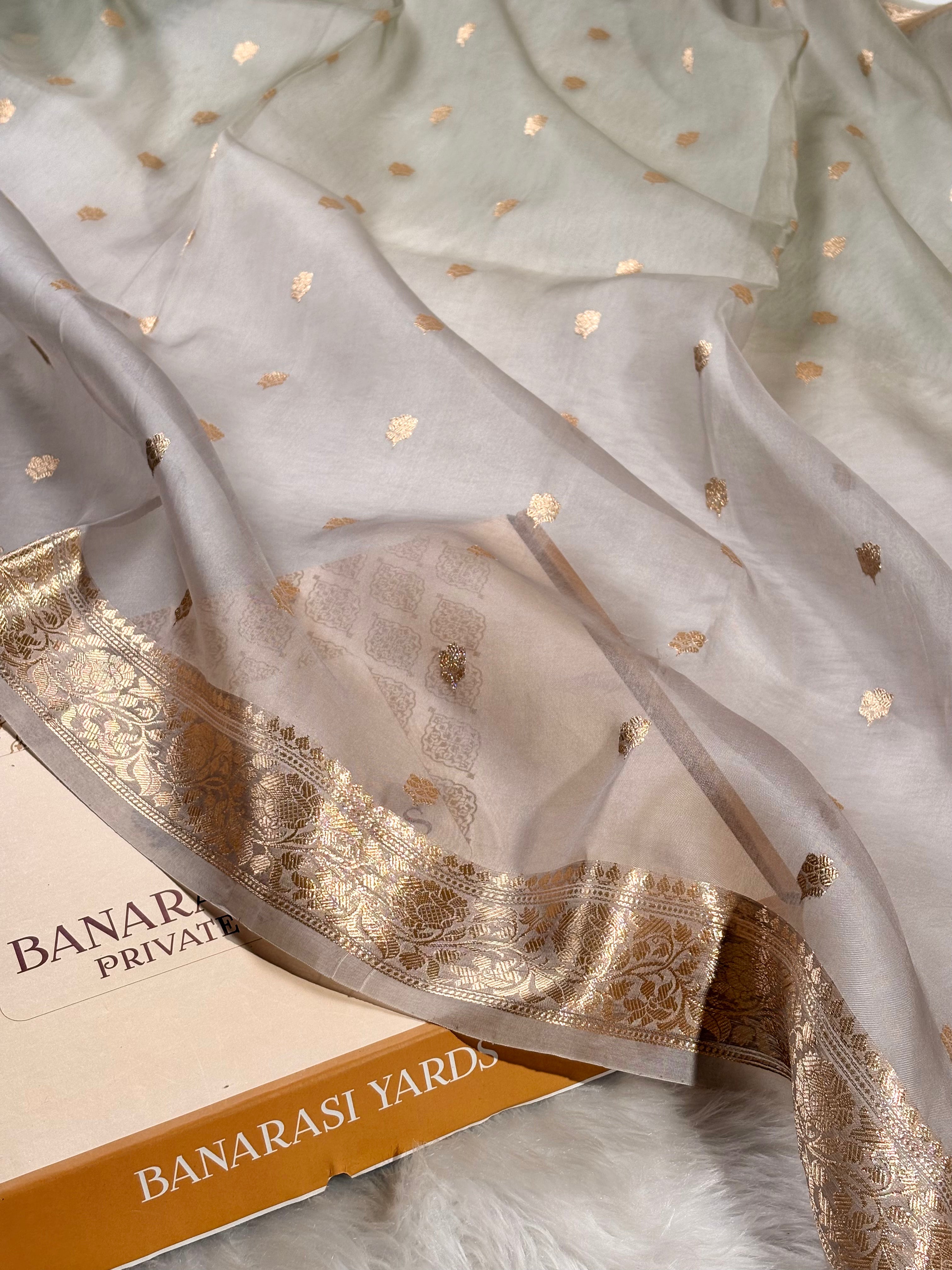 A sheer off-white organza saree with scattered gold foil motifs and a wide ornate gold zari border, draped over a box labeled Banarsi Yards.