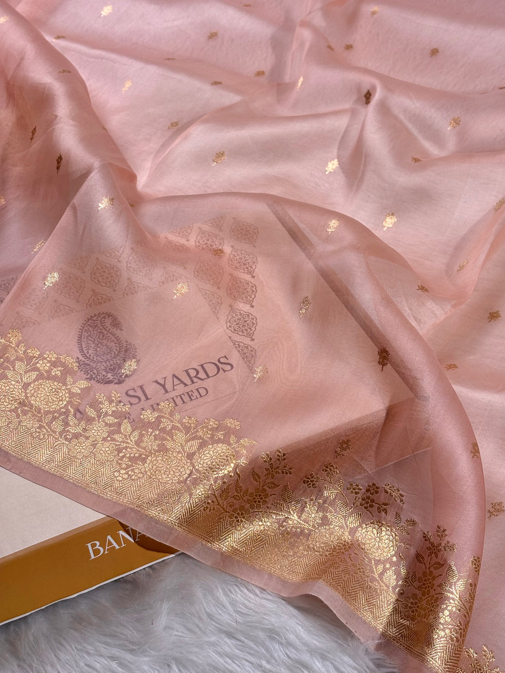 A draped light pink kora organza saree with a shimmering gold floral border and scattered gold motifs, shown on a white faux-fur surface with part of a branded box visible.