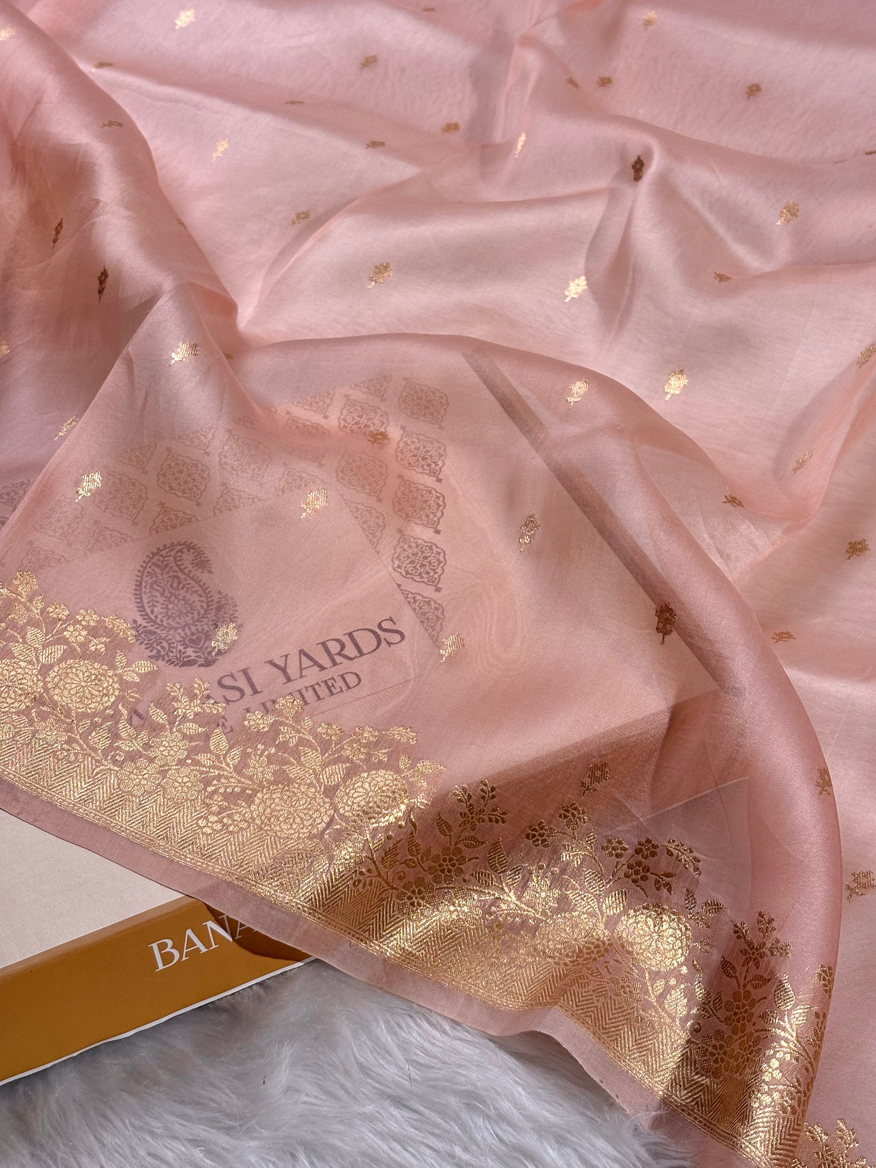 A draped light pink kora organza saree with a shimmering gold floral border and scattered gold motifs, shown on a white faux-fur surface with part of a branded box visible.