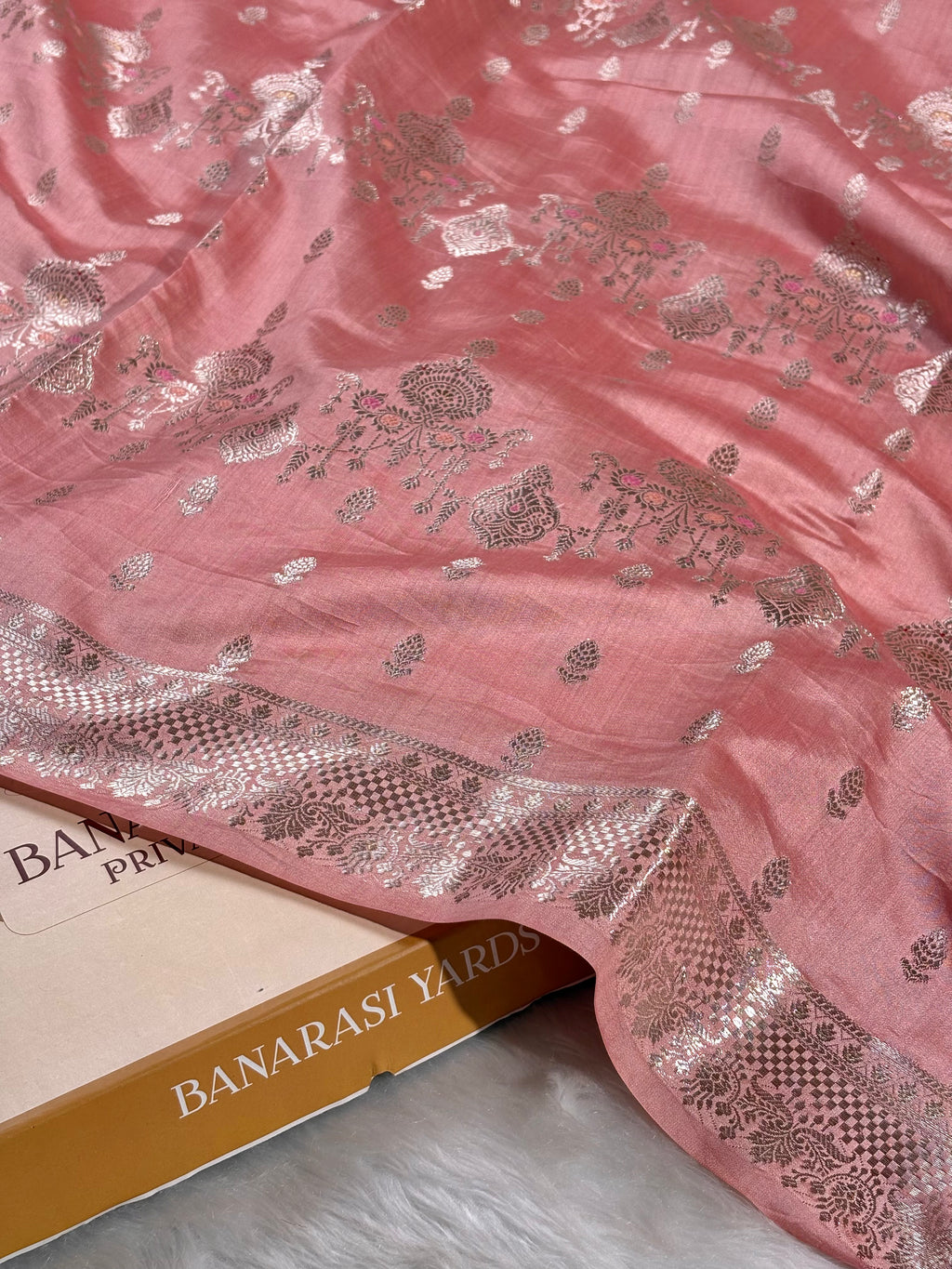 A pink Banarasi silk saree with silver floral zari weaving and a wide decorative border, draped over a box labeled 'BANARASI YARDS'.
