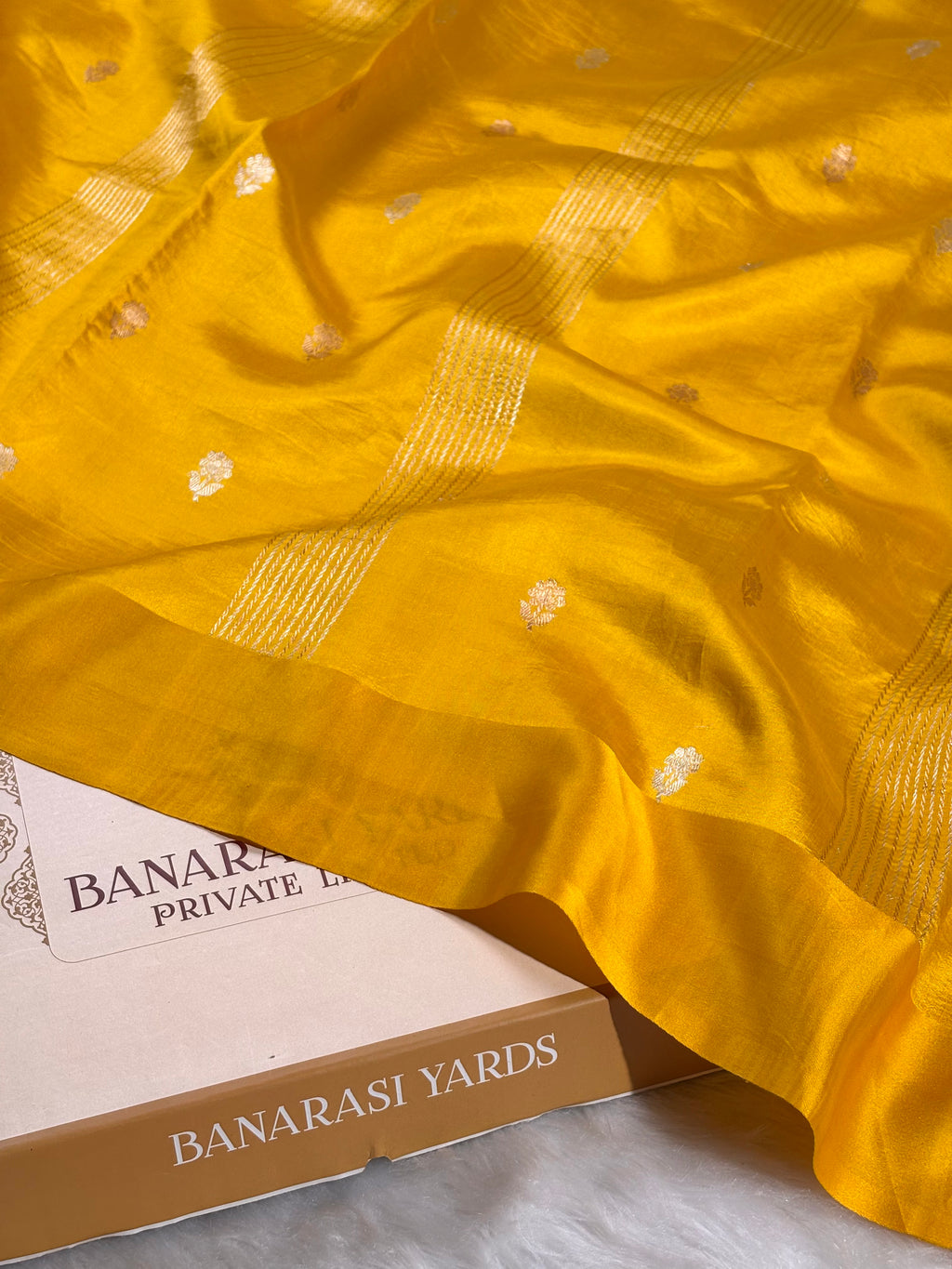 A close-up of a bright yellow Banarasi silk saree draped over a box labeled 'BANARASI YARDS'. The saree features subtle woven motifs and stripes, showcasing its texture and sheen.