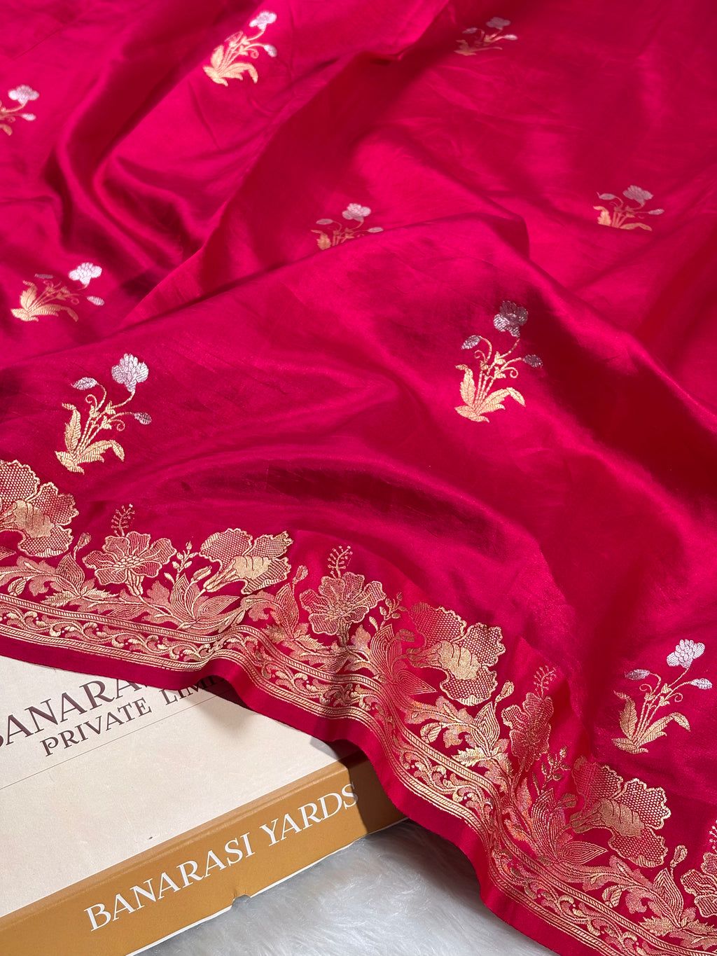 A close-up of a rich magenta-pink silk saree with intricate alfi mina floral weaving and a wide gold Banarasi border, draped elegantly with a Banarasi Yards box visible underneath.