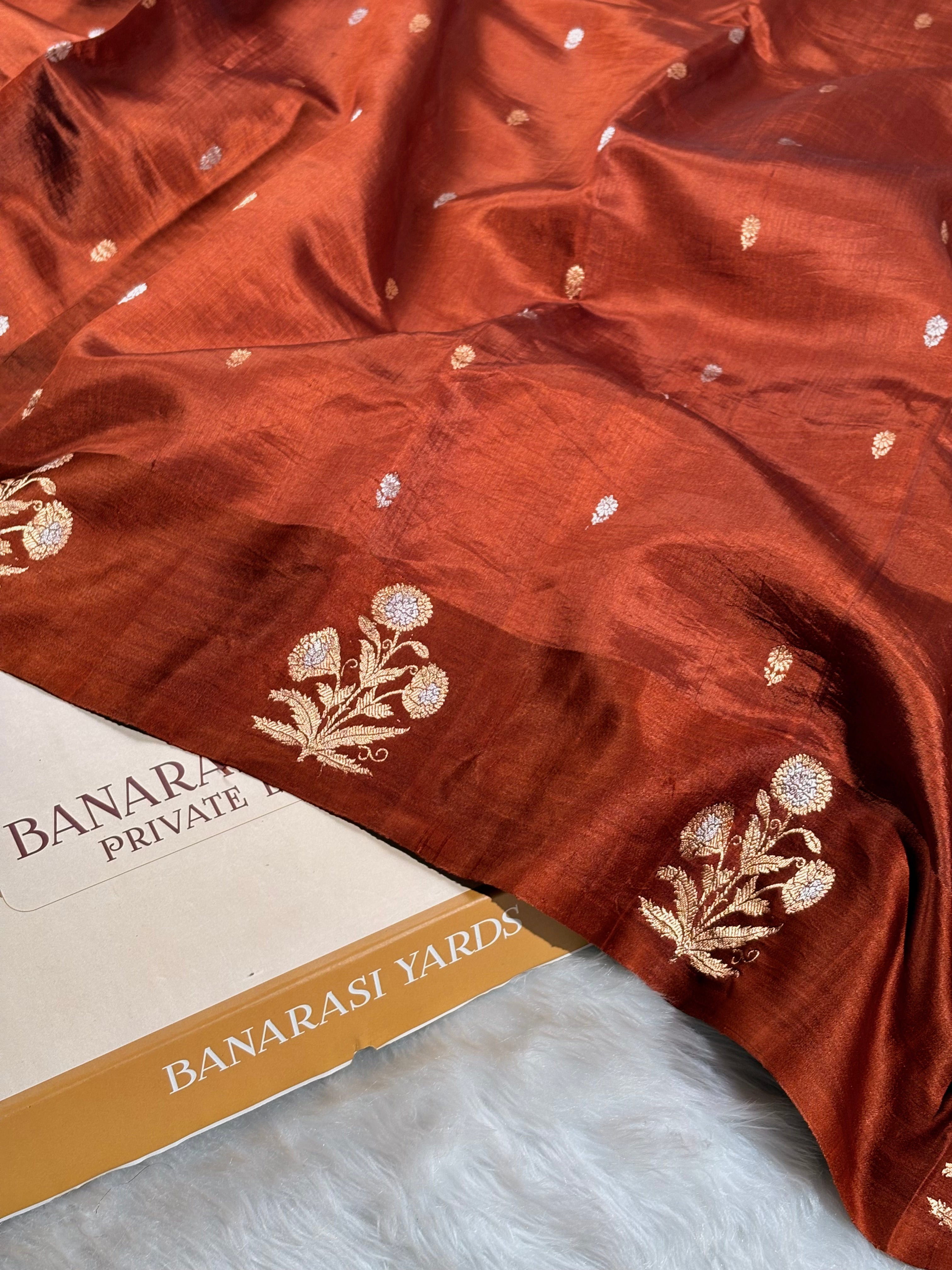 A rust-orange Banarasi silk saree with alfi zari floral weaving and small motifs, draped over a white faux fur surface next to a box labeled 'BANARASI YARDS'.