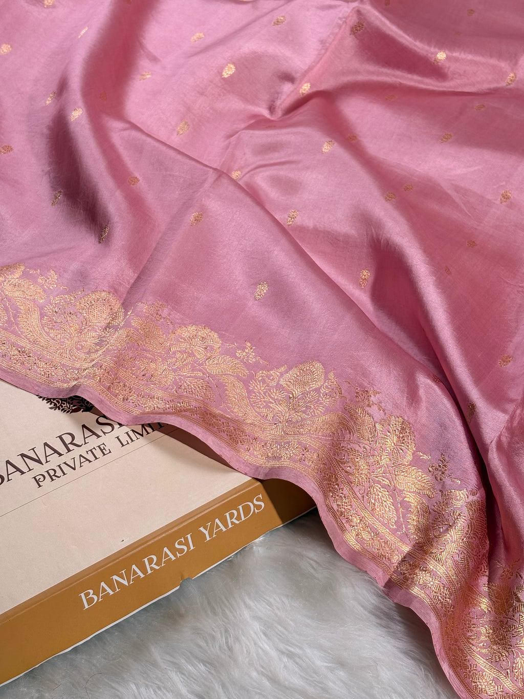 pure katan silk handloom Banarasi saree with a gold zari border and small gold motifs, draped over a box labeled 'BANARASI YARDS' on a white faux fur surface.