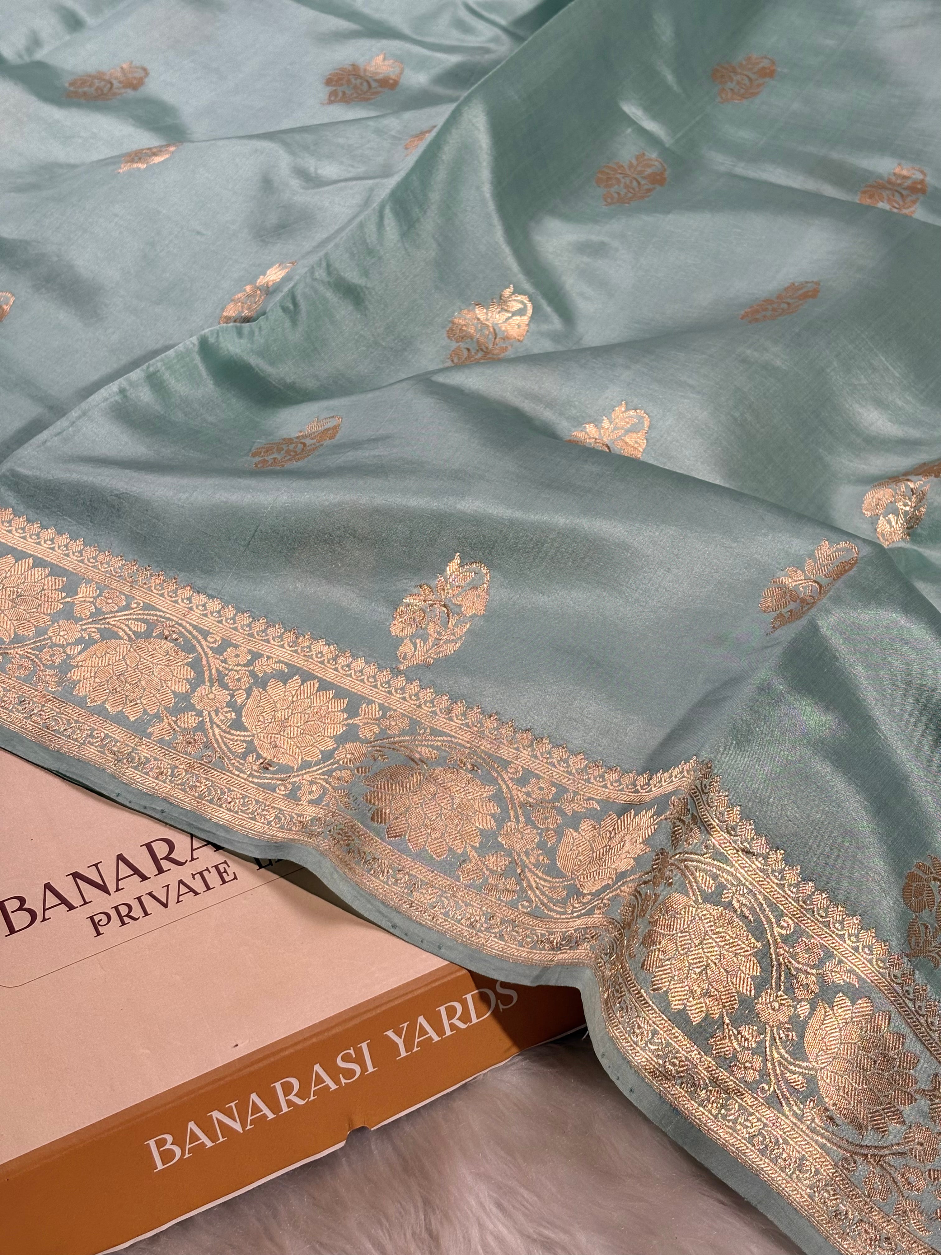 A sage green Banarasi silk saree with gold zari woven floral motifs and a wide ornate border, draped over a surface next to a cardboard box labeled 'BANARASI YARDS'.