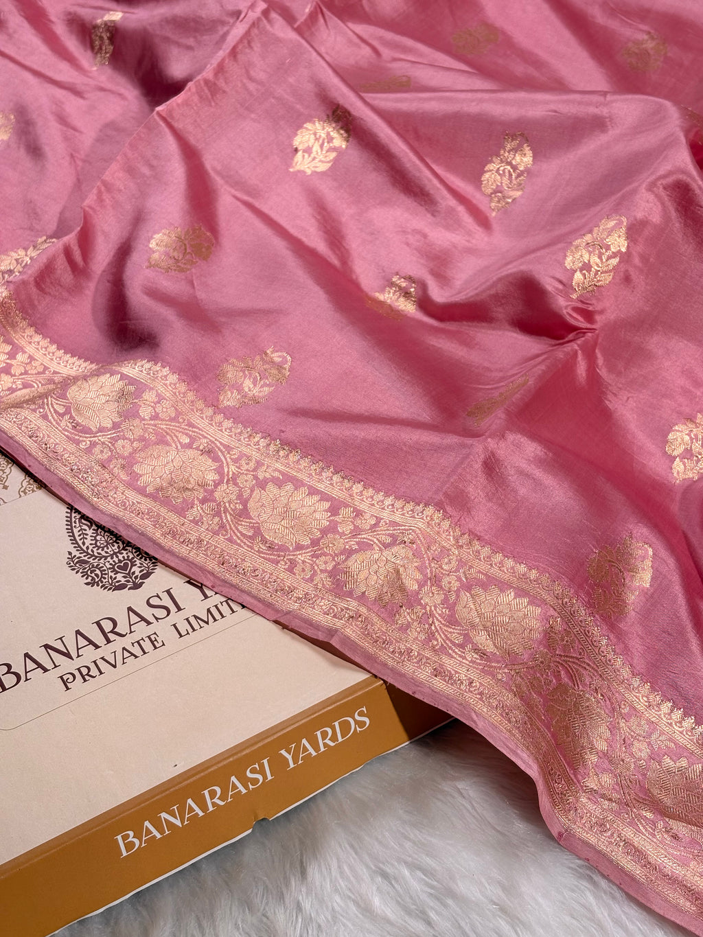 A pink Banarasi silk saree with gold zari floral motifs and a wide decorative border, draped over a box labeled 'BANARASI YARDS' on a white faux-fur surface.