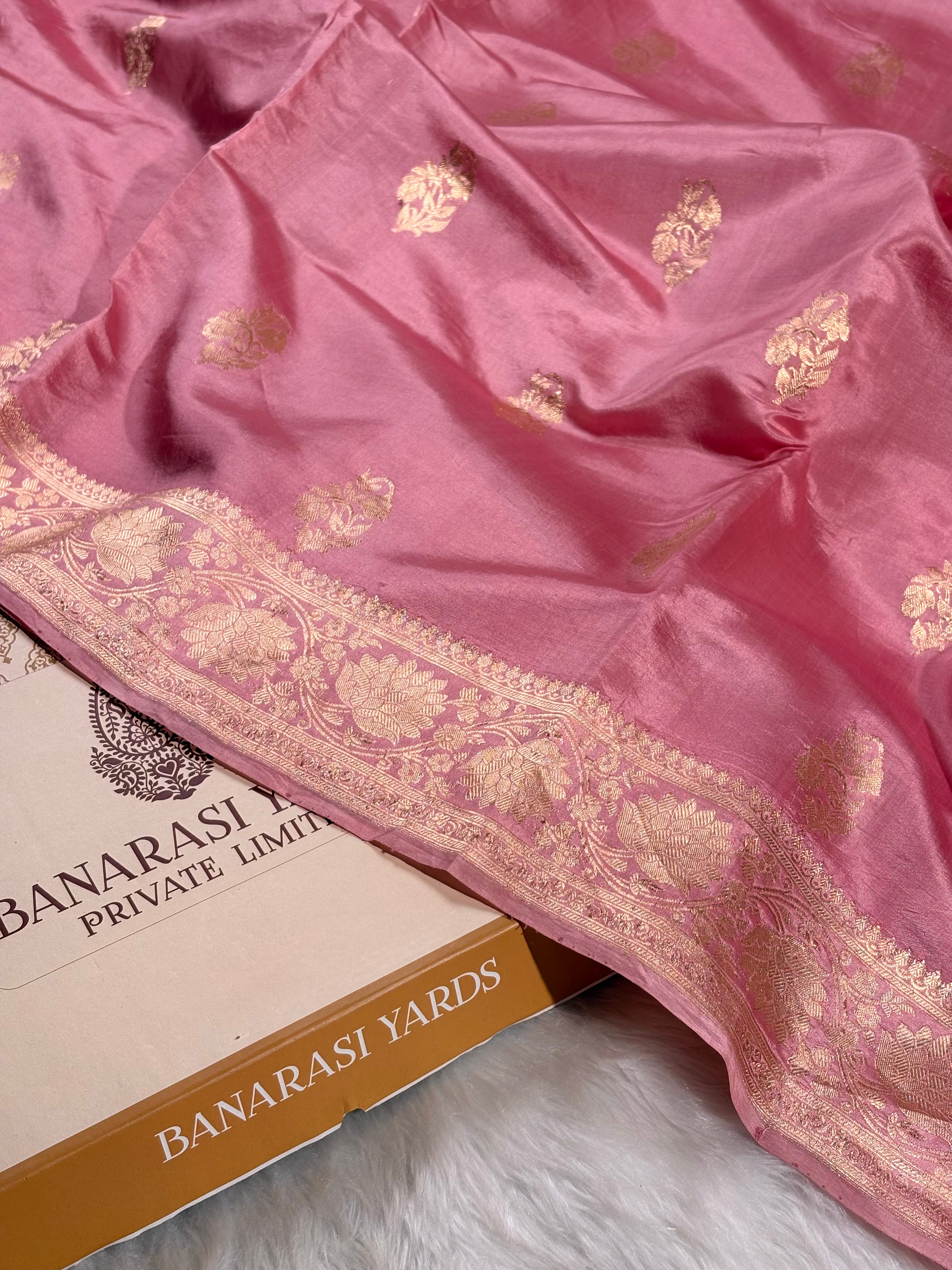 A pink Banarasi silk saree with gold zari floral motifs and a wide decorative border, draped over a box labeled 'BANARASI YARDS' on a white faux-fur surface.