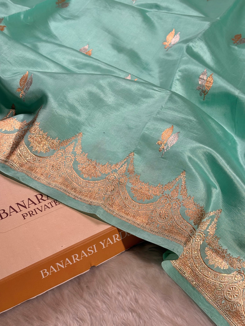 A mint green silk saree with intricate alfi mina zari weaving and a wide decorative border, draped over a brown Banarasi Yards box on a gray surface.