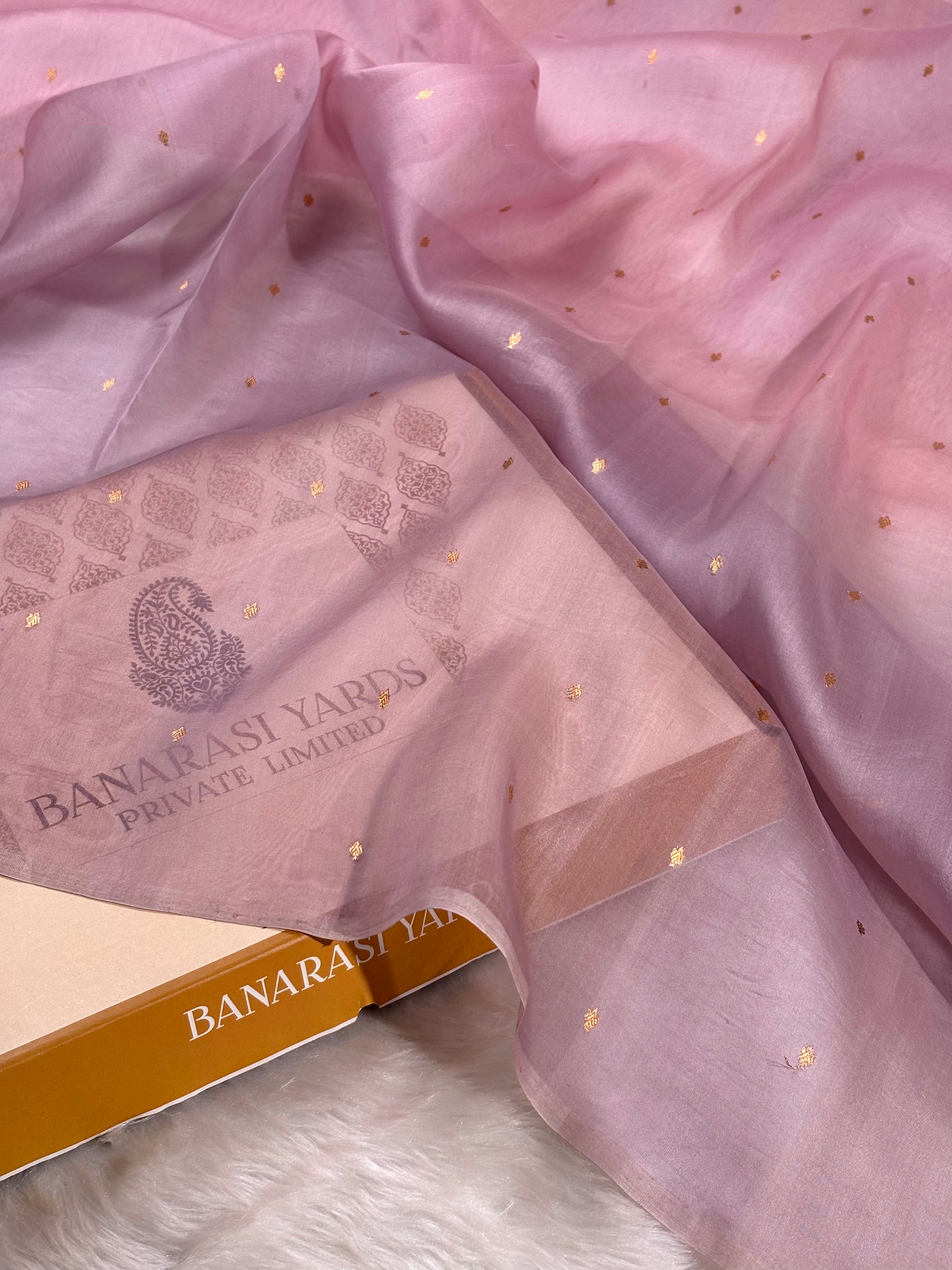 A draped pink pure kora organza saree with small gold foil star patterns, shown alongside a branded Banarasi Yards box on a white faux-fur surface.