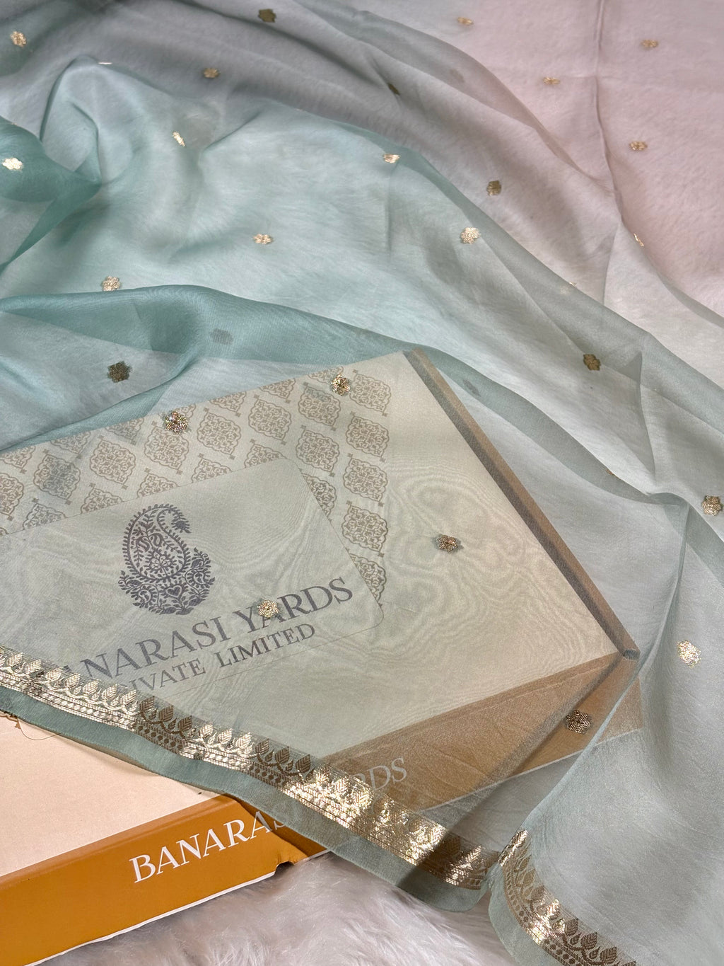 A pale mint green sheer saree with small gold buti weaving and a gold border, draped over a white Banarasi Yards Private Limited box.