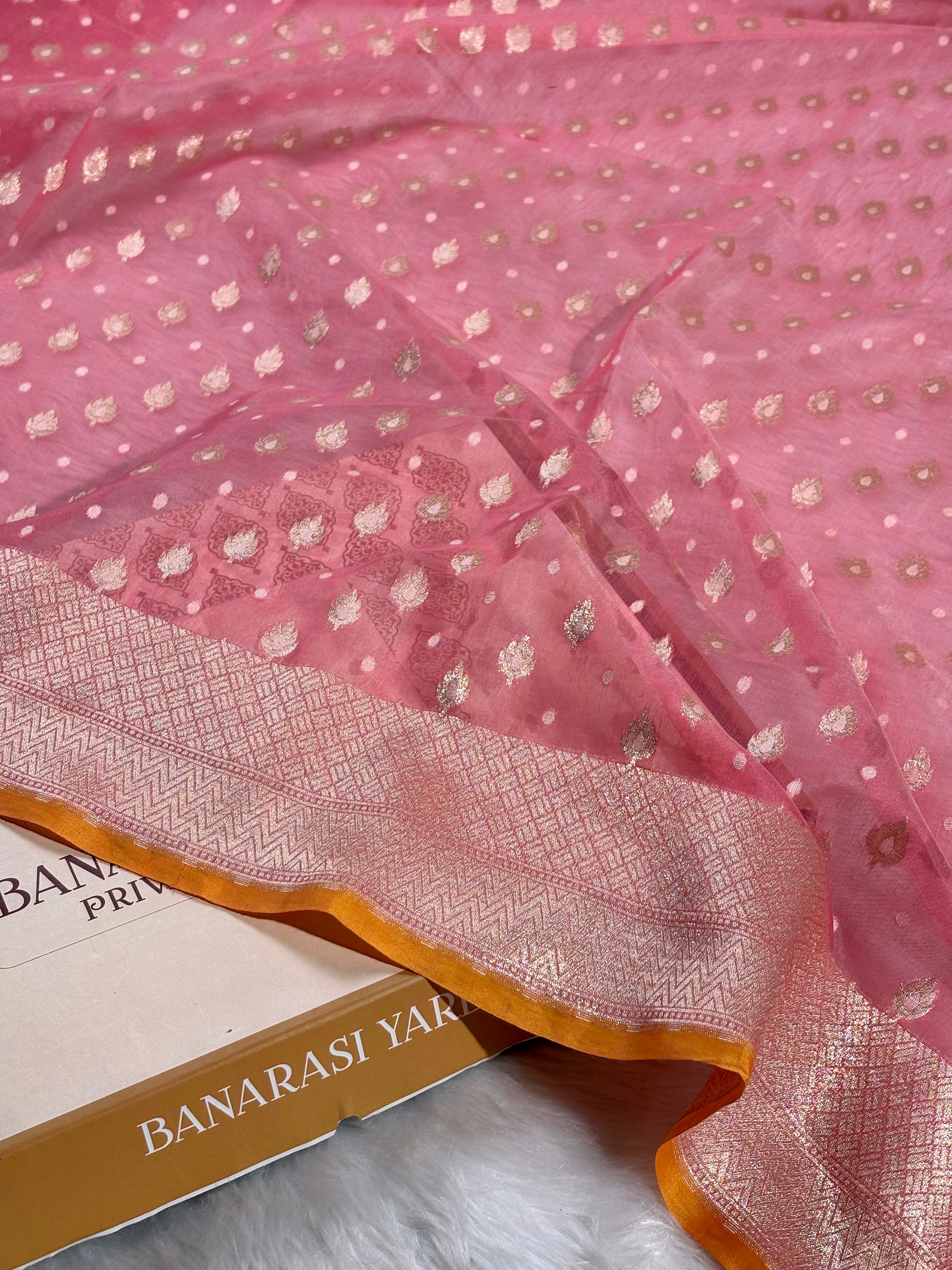 Pink saree with gold zari floral motifs and a wide gold border, draped over a Banarasi Yards box on a white faux fur surface.