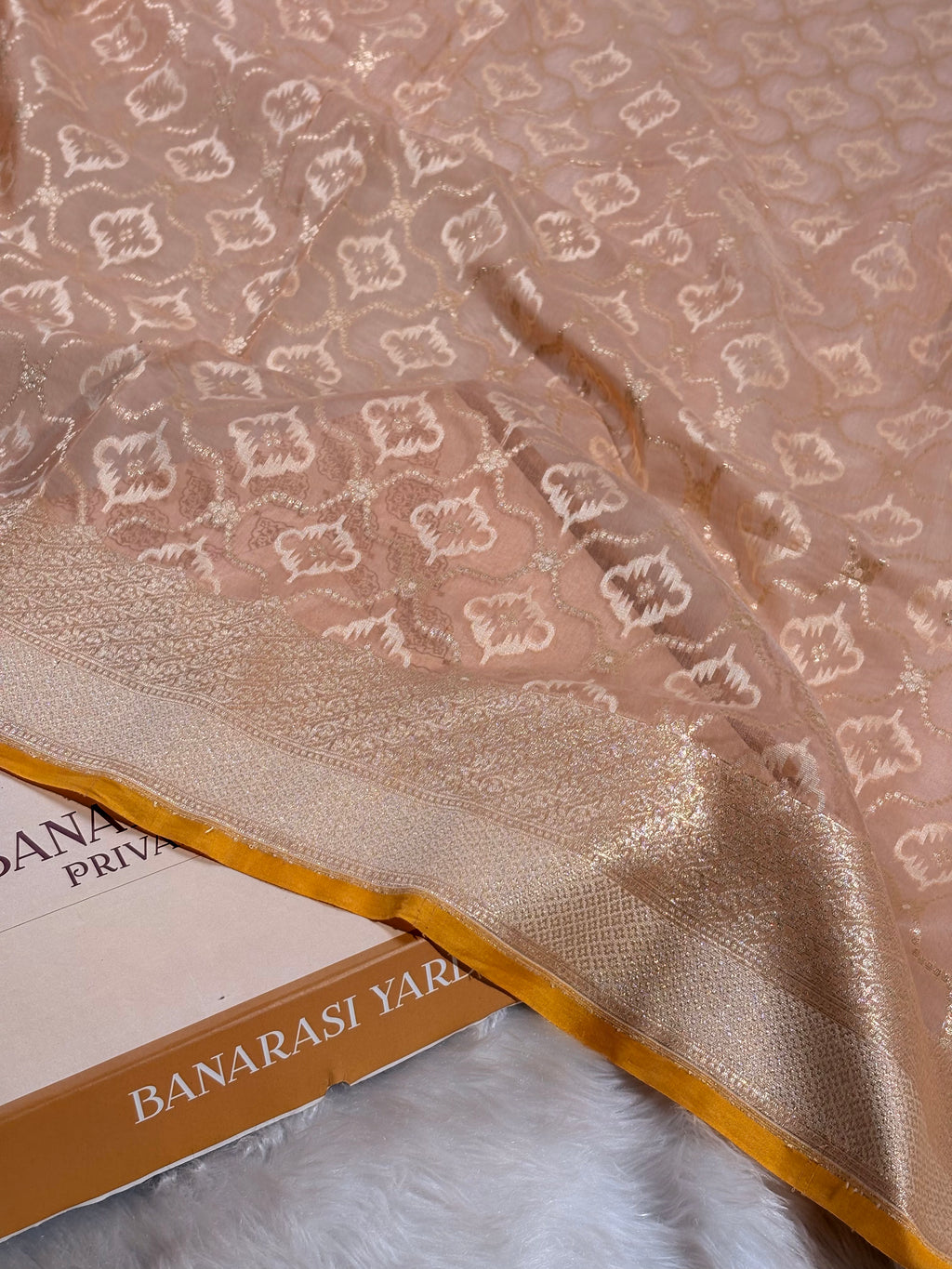 A close-up of a peach-beige Banarasi silk saree with intricate gold and white woven floral motifs and a wide gold border, draped over a boox labeled 'BANARASI YARDS' on a white faux fur surface.