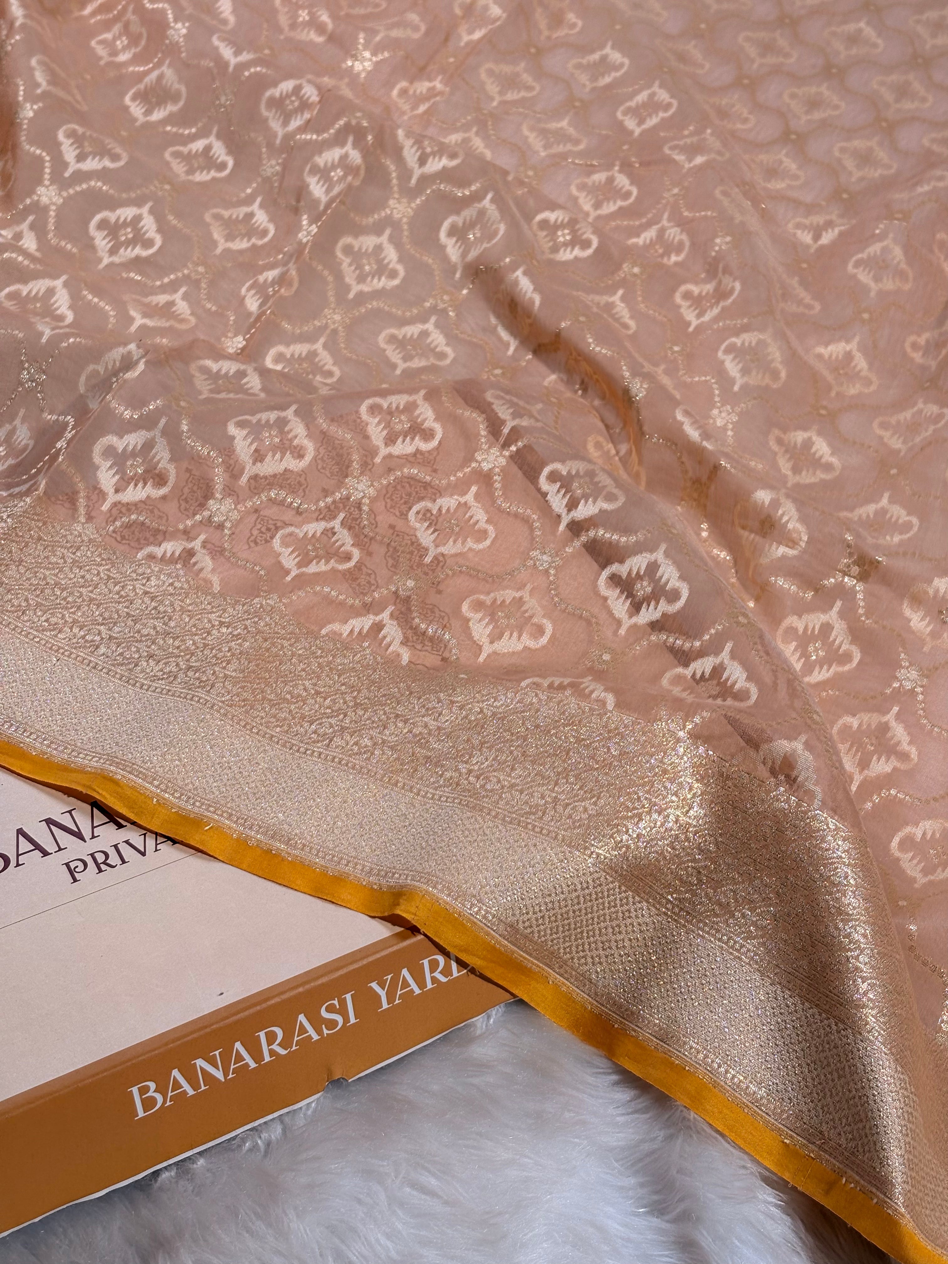 A close-up of a peach-beige Banarasi silk saree with intricate gold and white woven floral motifs and a wide gold border, draped over a boox labeled 'BANARASI YARDS' on a white faux fur surface.