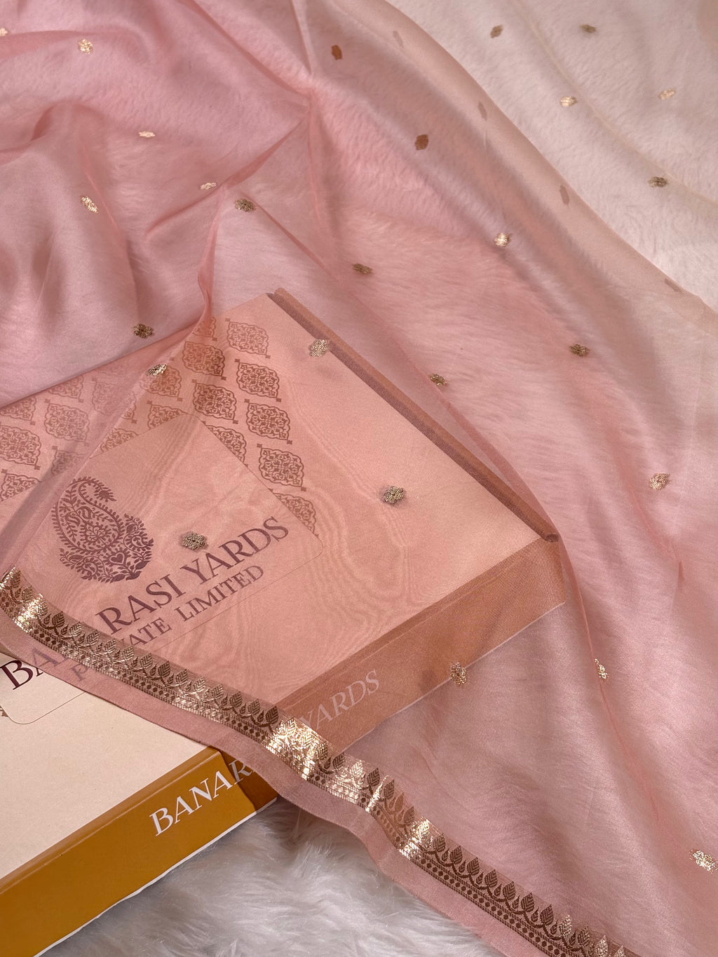 A close-up of a light pink saree, draped over a matching pink box. The saree features delicate gold zari weaving and small gold dots, with a gold zari border.