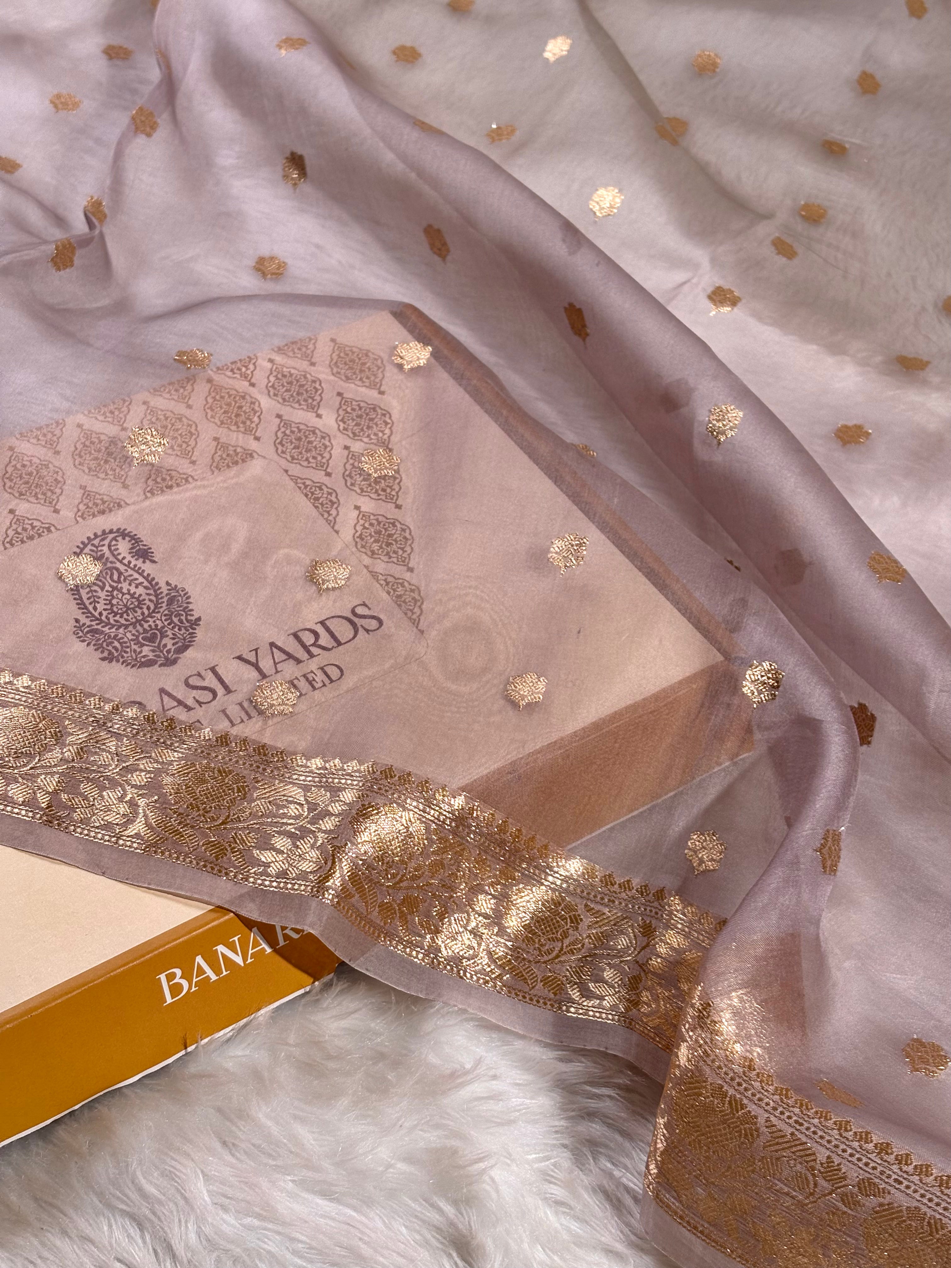 A close-up of a light blush pink organza saree with scattered gold foil motifs and a wide gold woven border, draped over a matching box. The box shows the brand name 'BANARASHI YARDS'.
