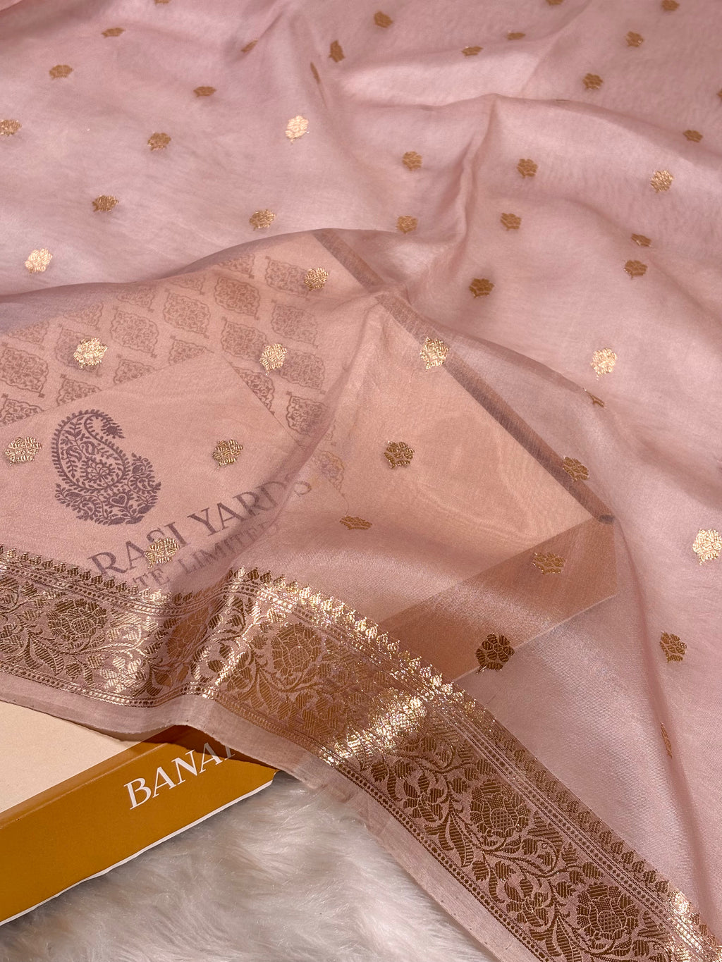 A dusty pink organza saree with gold foil motifs and a wide gold zari border, draped elegantly with gold-colored box partially visible.
