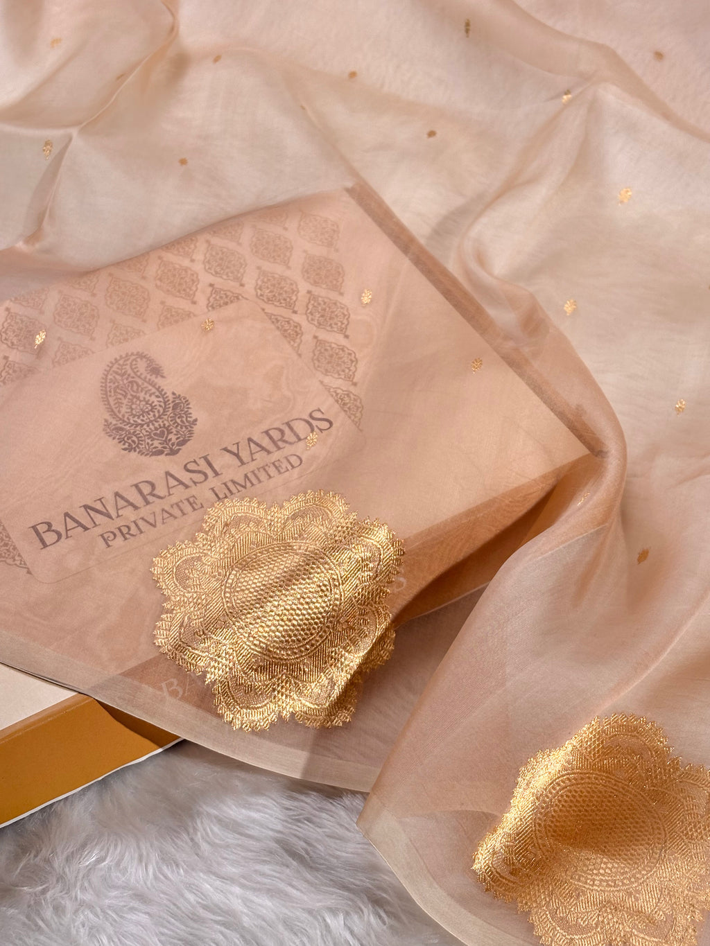 A peach-beige sheer organza with scattered gold foil dots, draped over a matching branded paper box printed with 'BANARASI YARDS PRIVATE LIMITED'.