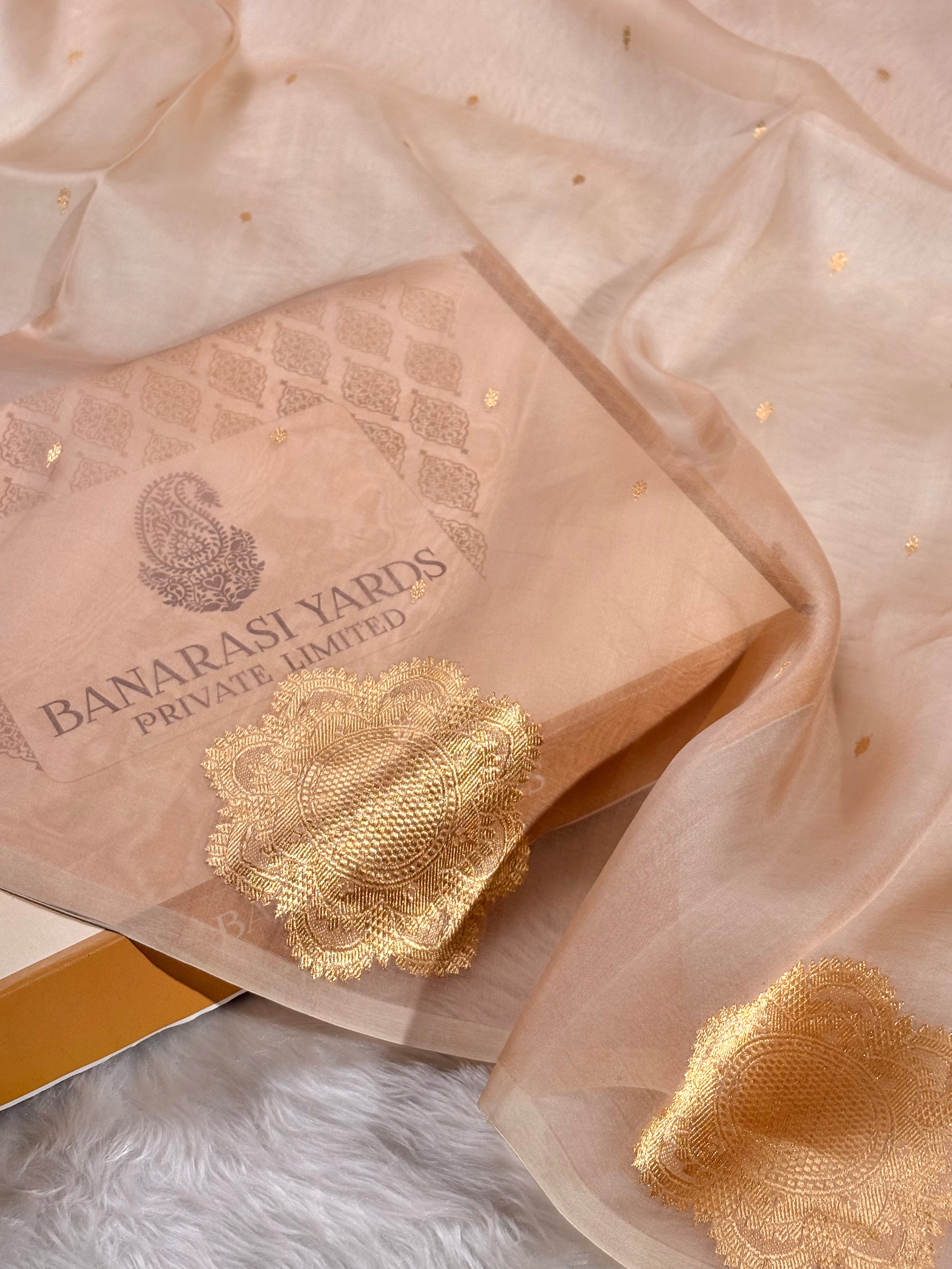 A peach-beige sheer organza with scattered gold foil dots, draped over a matching branded paper box printed with 'BANARASI YARDS PRIVATE LIMITED'.