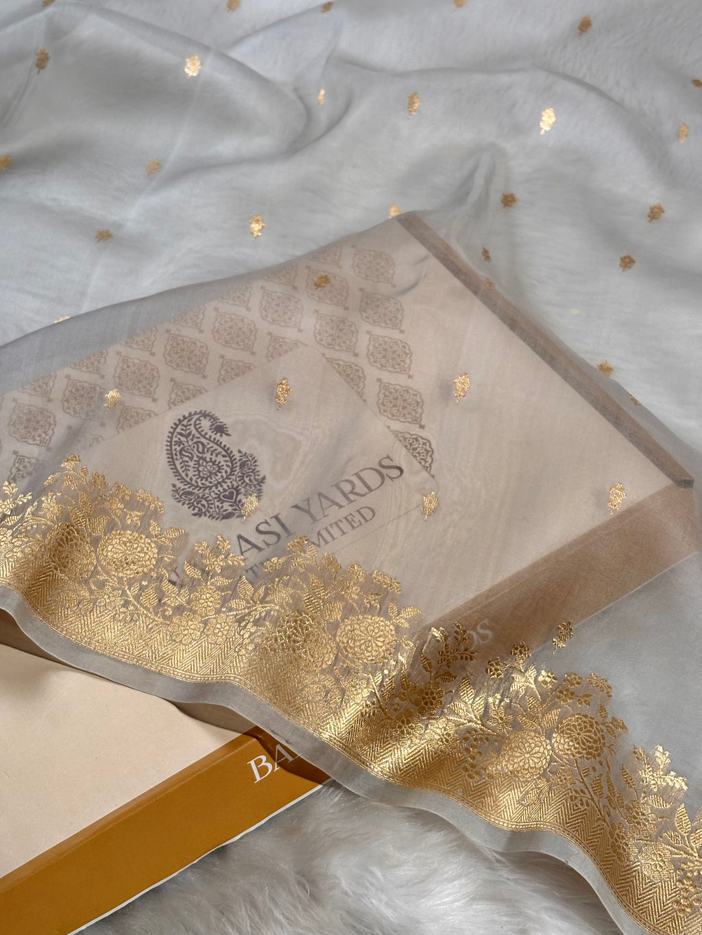 A close-up of a printed saree box with a decorative gold border, placed on a white textured surface with a matching white fabric backdrop dotted with small gold flecks. The brand name 'BANARASI YARDS' is visible on the box.