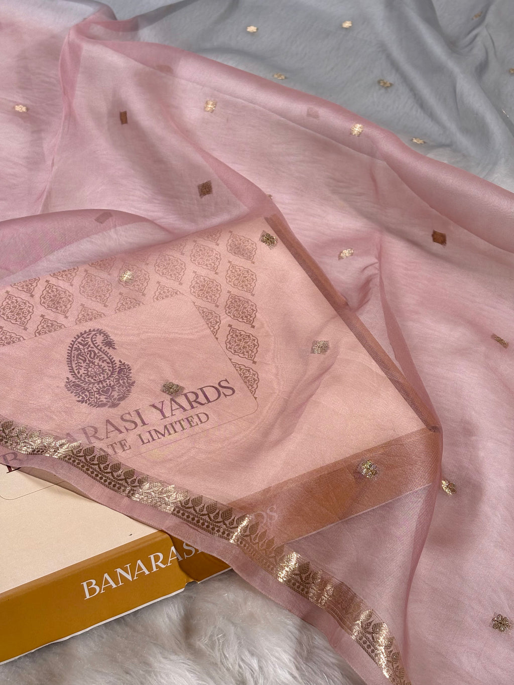 A light pink organza saree with a gold zari border and small gold foil dots. The inner lining shows the brand name 'Banarasi Yards'.