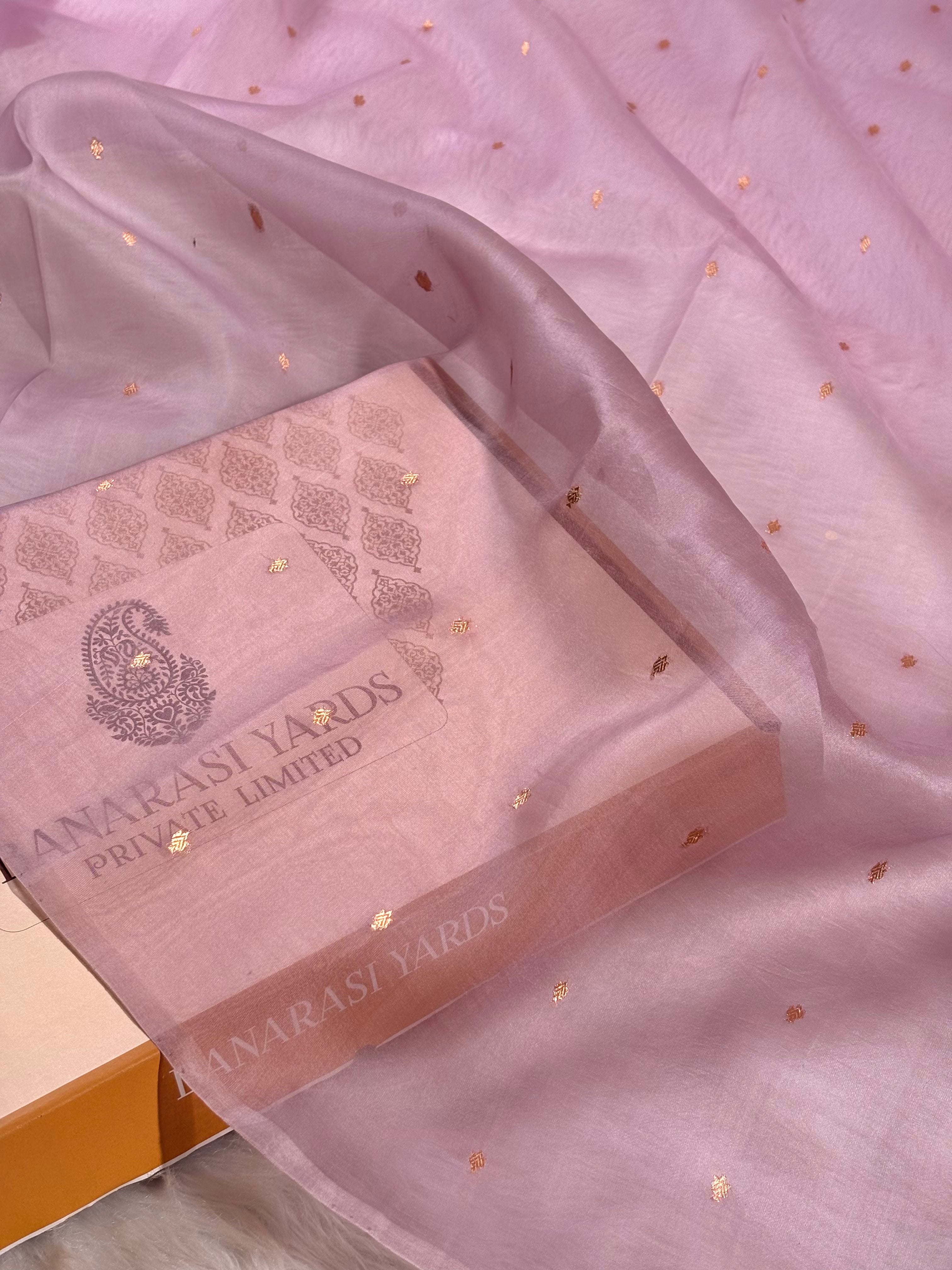 A close-up of a pink kora organza saree with scattered small gold foil motifs, draped over a matching pink branded box labeled 'Banarasi Yards Private Limited'.