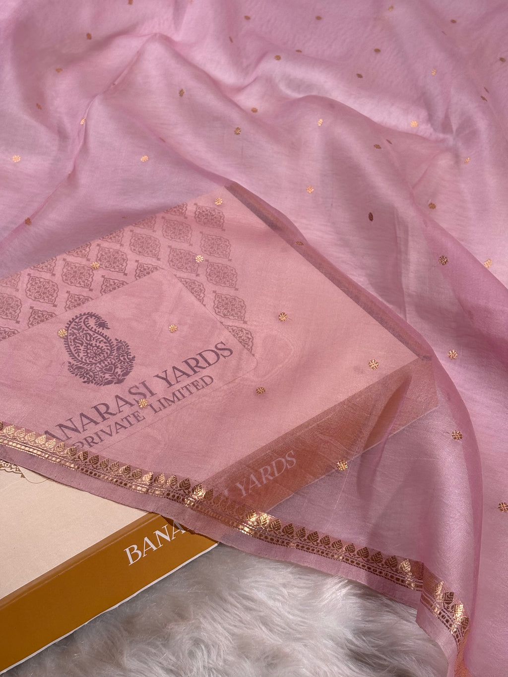 A light pink kora organza saree with scattered gold foil dots and a decorative border, draped over its matching pink packaging box featuring the 'Banarasi Yards' logo.