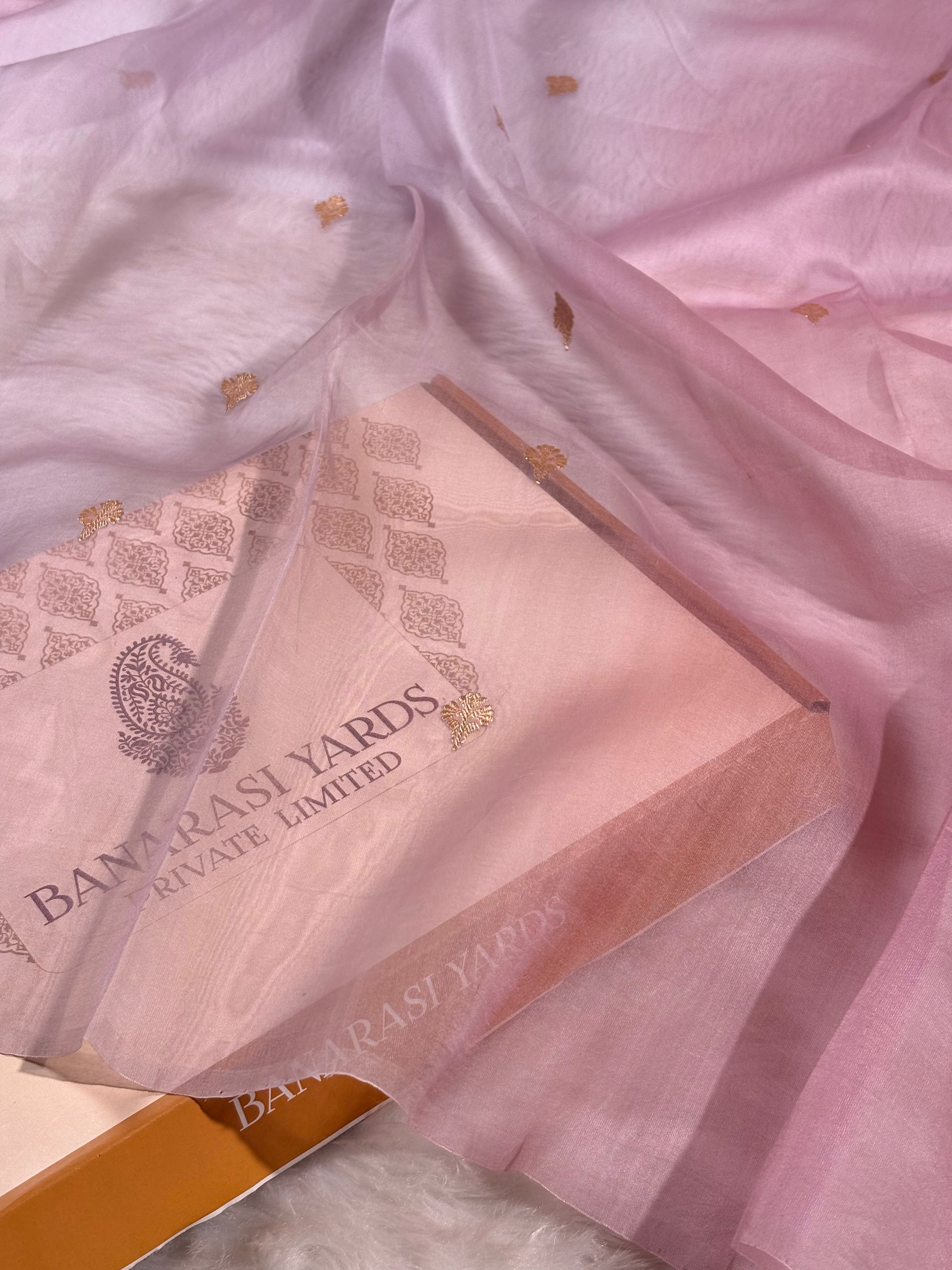 A close-up of a light pink kora organza Banarasi saree with small gold motifs, draped over a matching pink gift box printed with 'BANARASI YARDS PRIVATE LIMITED'.