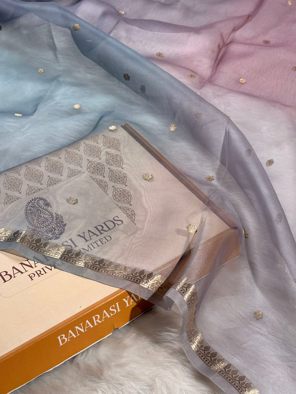 A kora organza Banarasi saree with a soft ombré transition from light blue to pale pink, decorated with small gold foil dots and a gold border, draped over a Banarasi yards sample box.