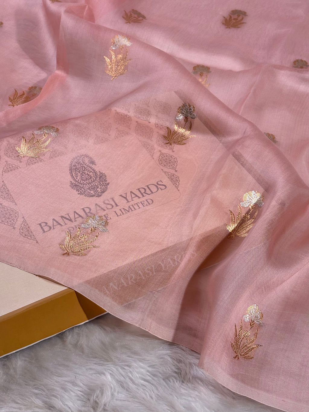 A draped light pink organza saree with alfi mina buta floral weaving,styled on a white faux fur surface, with box.