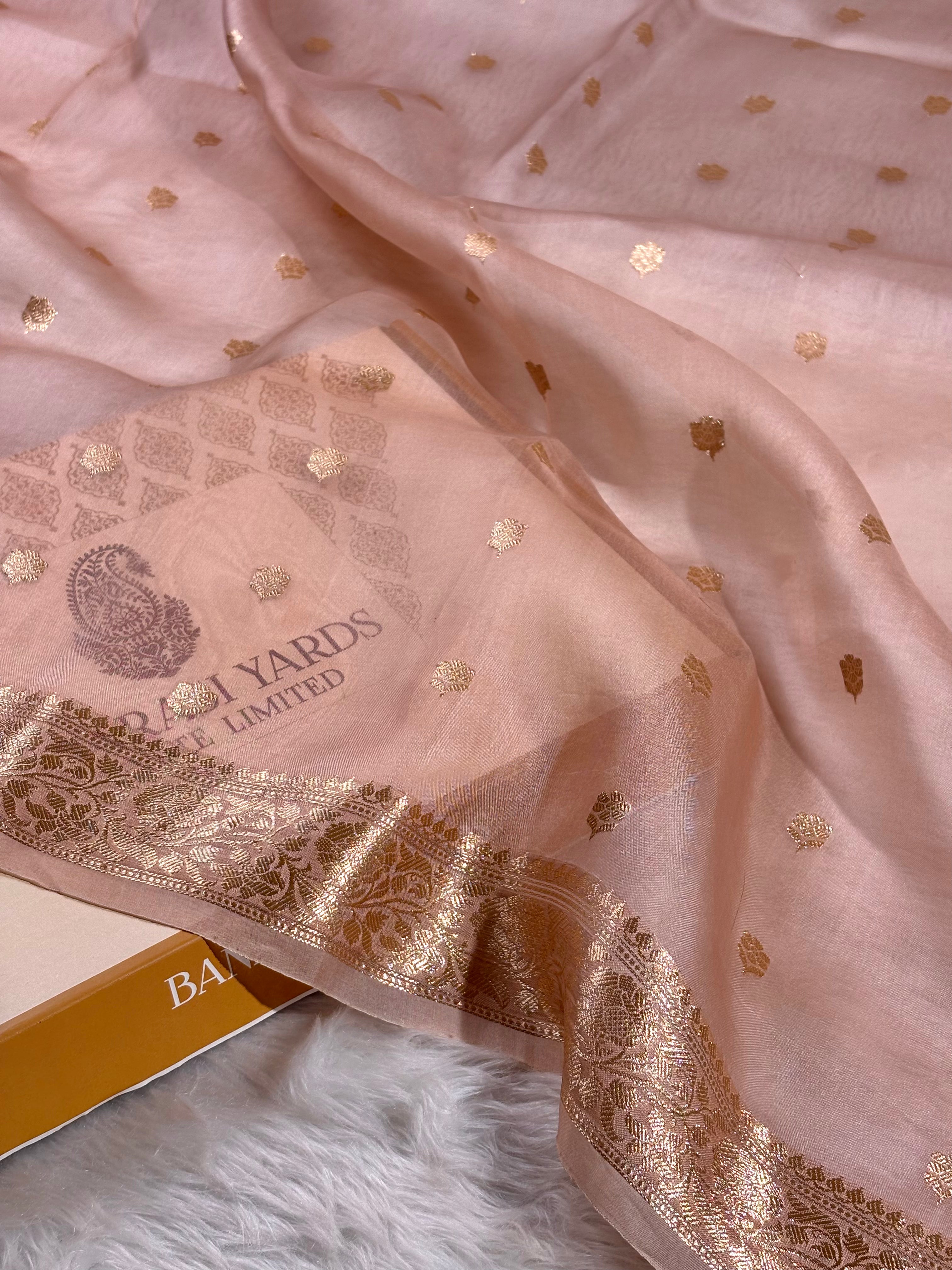 A draped light pink organza saree with scattered gold foil motifs and a wide ornate gold zari border, shown on a white faux-fur surface with part of the brand box visible.