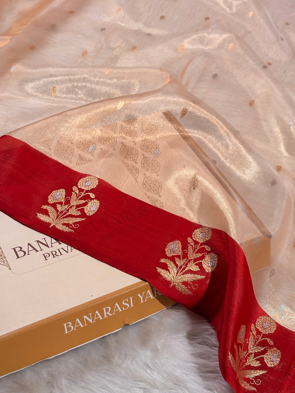 A close-up of a Banarasi silk saree draped over a box. The saree is a rich red satin border with intricate gold floral weaving.